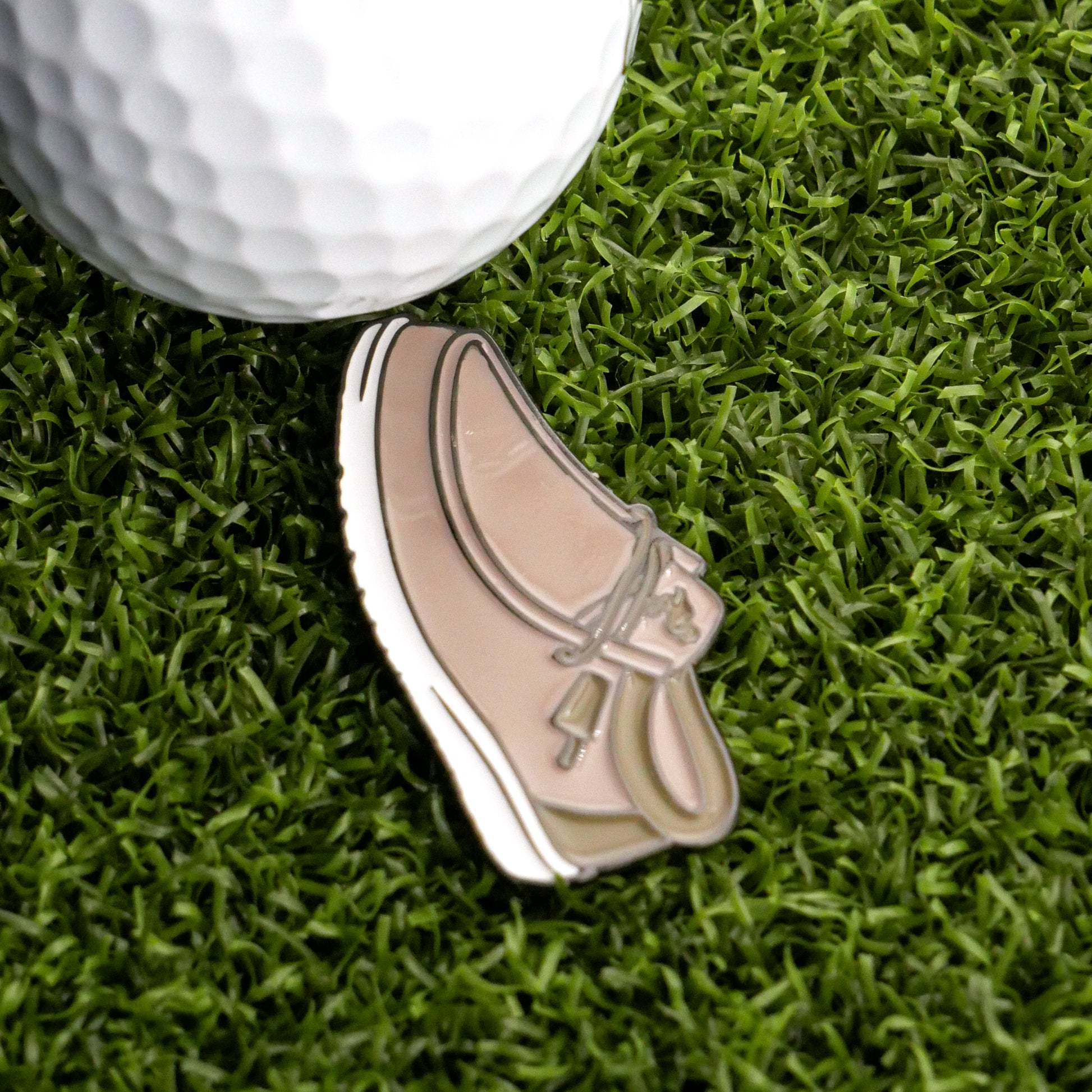 A close-up of a golf ball on artificial grass next to the Ballmarkers.com Yo Guy Slip-On Loafer Ball Marker, shaped like a comfortable shoe.