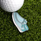 The Yo Guy Slip-On Loafer Ball Marker by Ballmarkers.com is a light blue, shoe-shaped pin resting on green artificial grass beside a white golf ball, serving as a unique and comfortable ball marker.