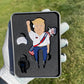 A white-gloved hand holds a Pin Creatures TRUMP Divot Tool box. The packaging shows a cartoon U.S. president figurine with a USA sash, American flag golf club, MAGA badge, and divot tool golf ball marker. Grass is visible in the background.