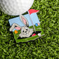 A golf ball sits on artificial grass beside the Ballmarkers.com Easter Bunny Egg Hunt Ball Marker, featuring a smiling bunny peeking from a hole with a red flag and vibrant Easter eggs—a delightful golfer's accessory.