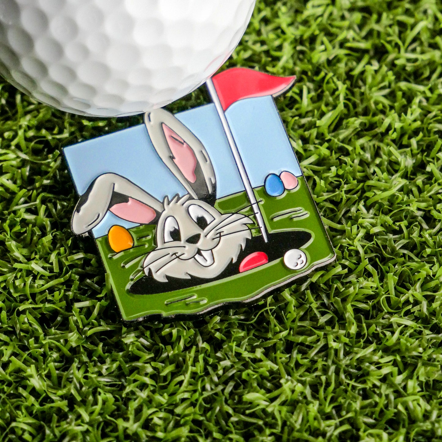 A golf ball sits on artificial grass beside the Ballmarkers.com Easter Bunny Egg Hunt Ball Marker, featuring a smiling bunny peeking from a hole with a red flag and vibrant Easter eggs—a delightful golfer's accessory.