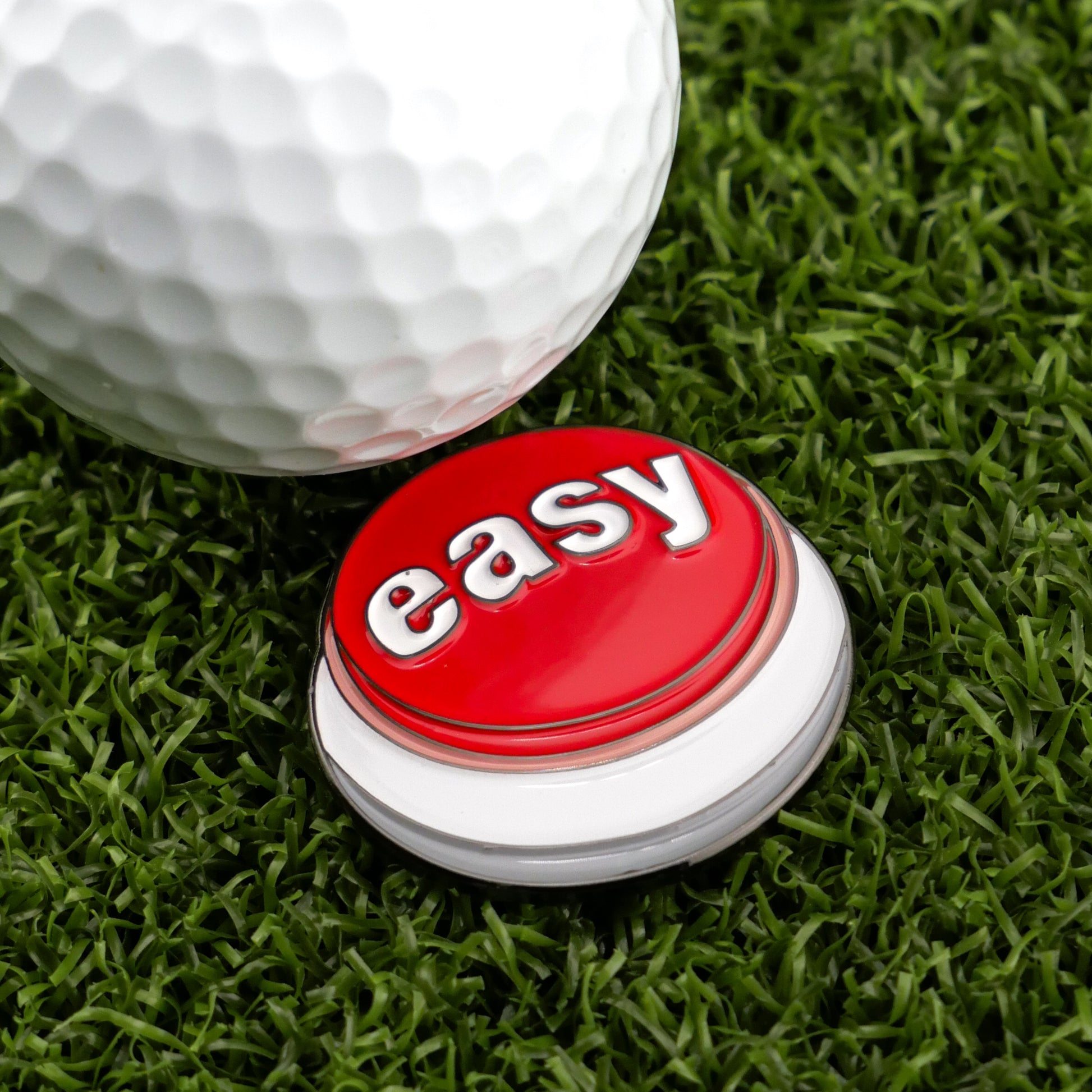 A close-up of a white golf ball on artificial green grass beside Logie Joe's Golf Easy Button, showcasing the appeal of personalized golf accessories like custom golf ball markers.