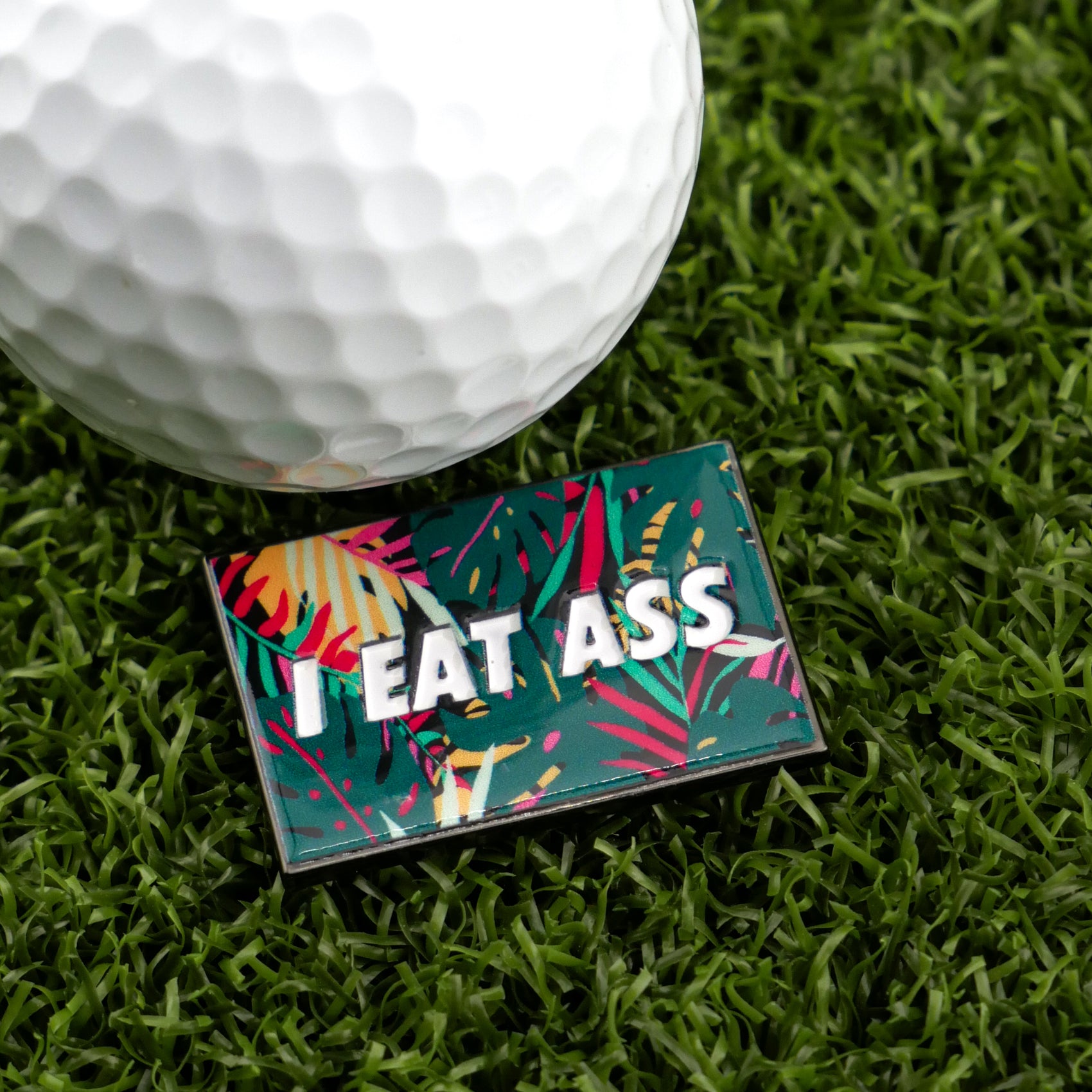 A close-up of a golf ball on artificial grass next to Logie Joe's Golf "I Eat *ss Tropical" pin—a bold and colorful accessory that's sure to add humor to your golf collection.
