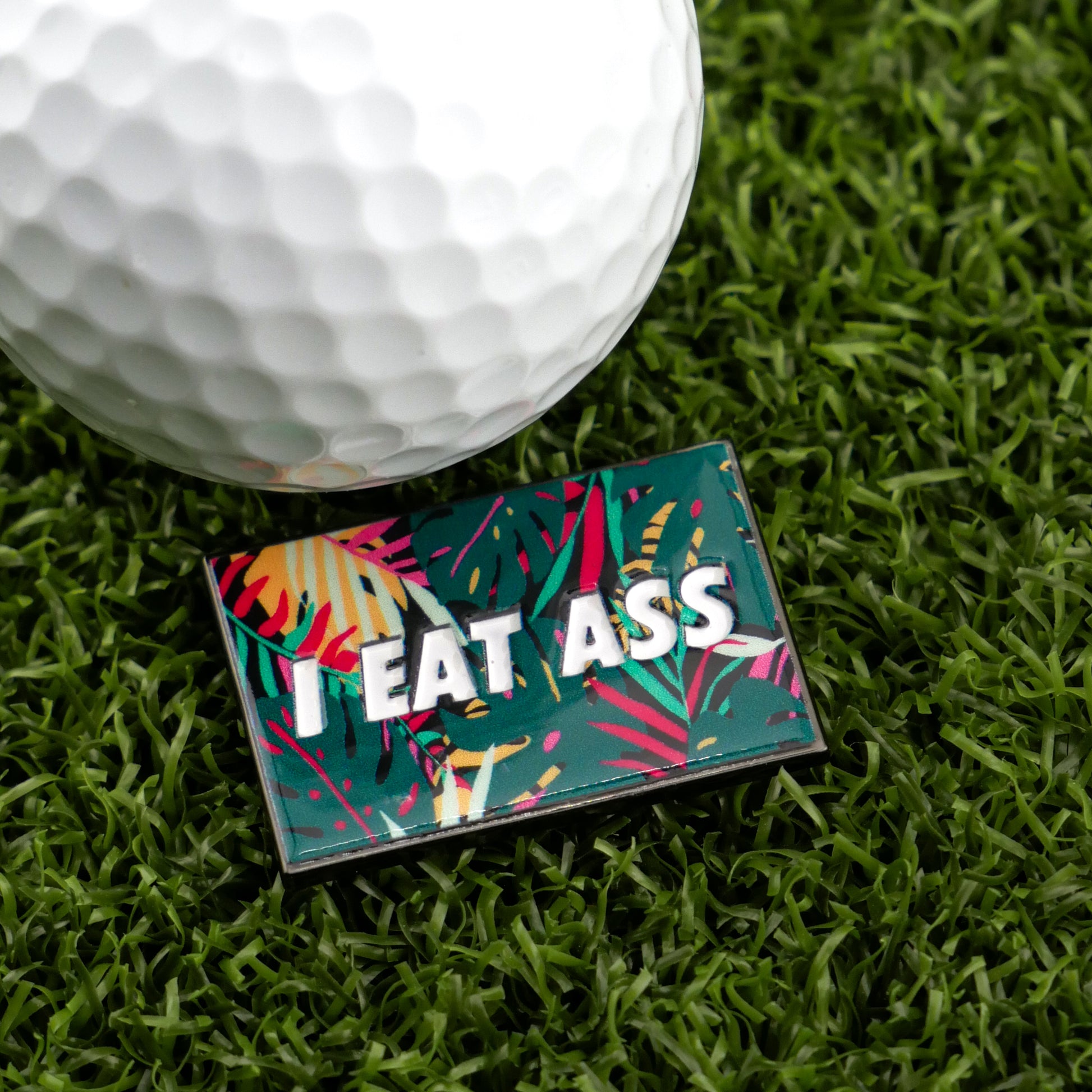 A close-up of a golf ball on artificial grass next to Logie Joe's Golf "I Eat *ss Tropical" pin—a bold and colorful accessory that's sure to add humor to your golf collection.