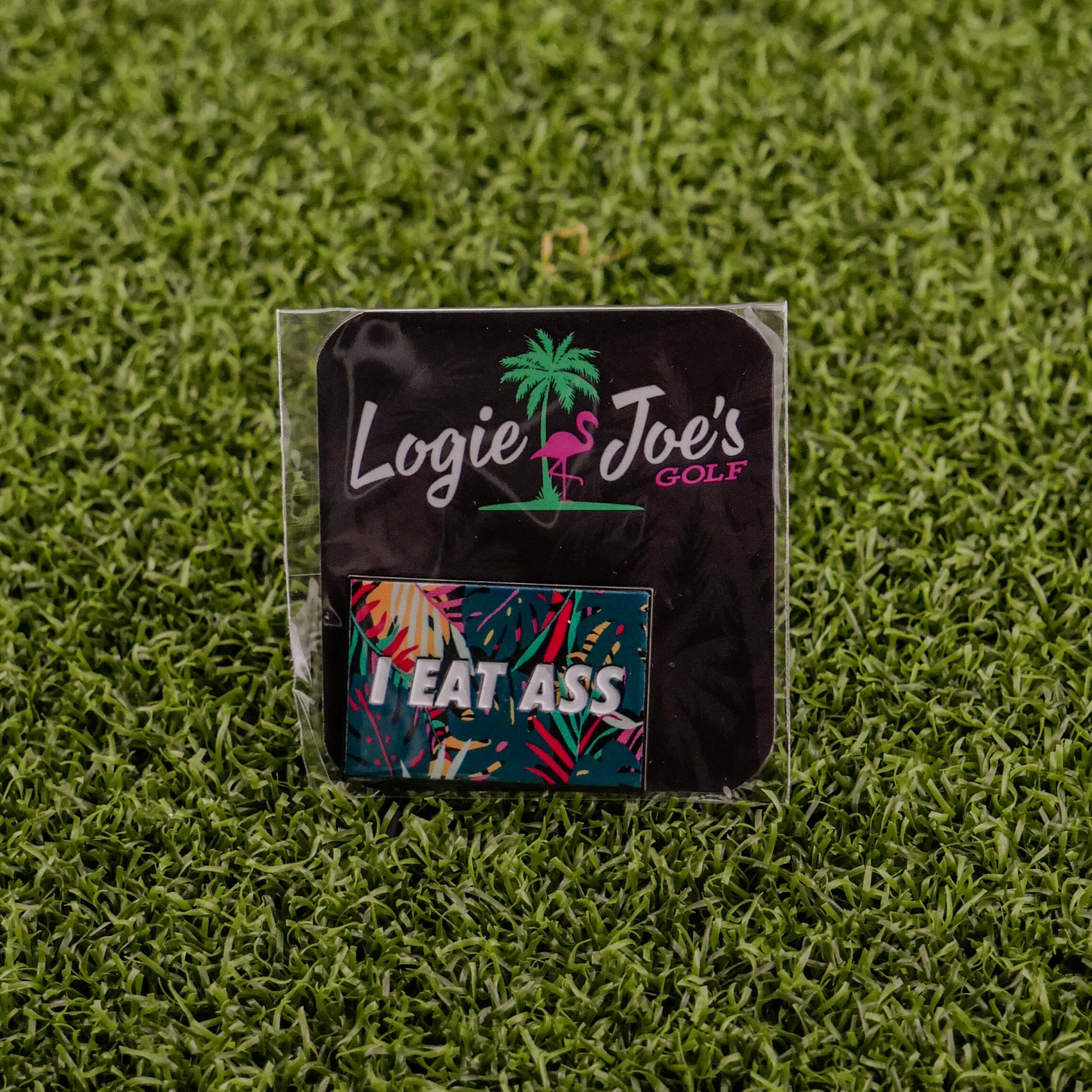 A small Logie Joe's Golf package featuring the "I Eat *ss Tropical" label and a colorful sticker sits on green turf—ideal for golf fans seeking funny accessories like a quirky divot tool or ball marker.