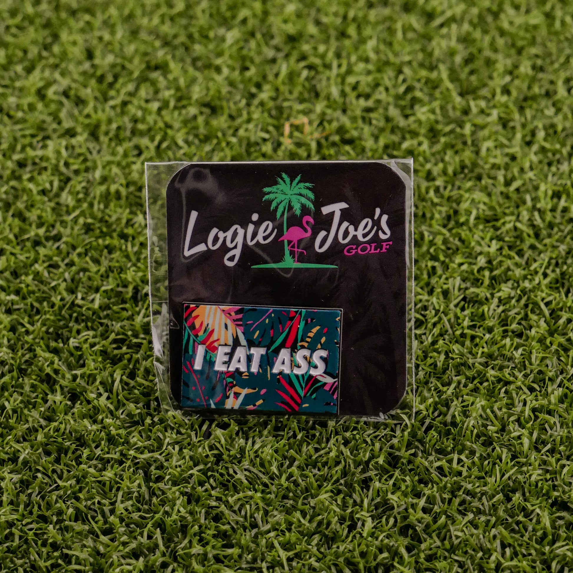 A small Logie Joe's Golf package featuring the "I Eat *ss Tropical" label and a colorful sticker sits on green turf—ideal for golf fans seeking funny accessories like a quirky divot tool or ball marker.