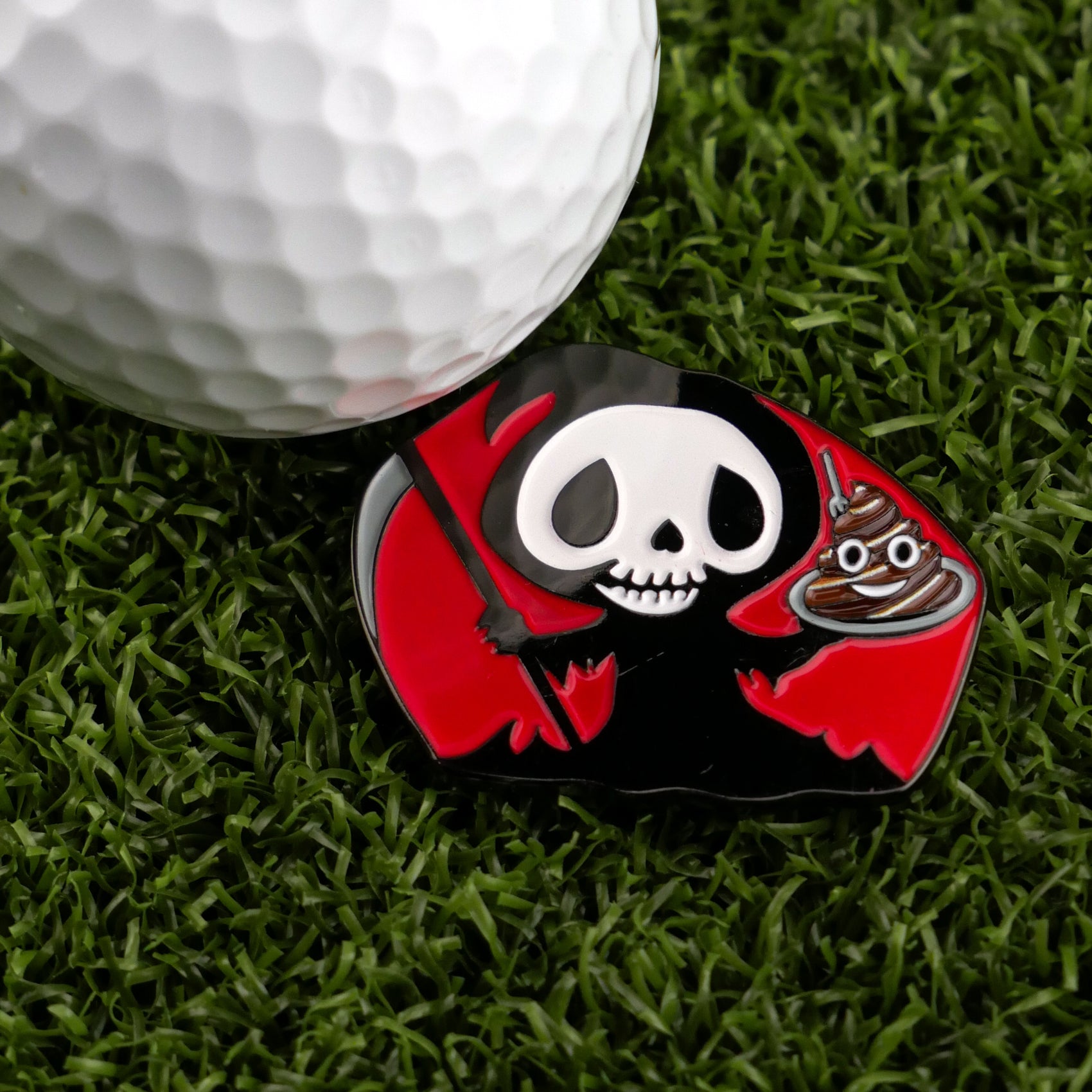On green grass, a golf ball rests beside Logie Joe's Golf "Eat Sh*t & Die" premium ball marker, featuring a cartoon grim reaper with a staff and poop emoji—a playful, artistic golf accessory.