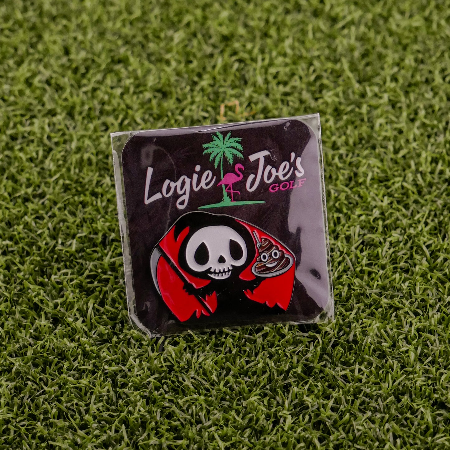 A premium Eat Sh*t & Die ball marker pin by Ballmarkers by Logie Joes, featuring a cartoon grim reaper with a golf club and Logie Joe’s Golf in white and pink, displayed on artificial green grass.