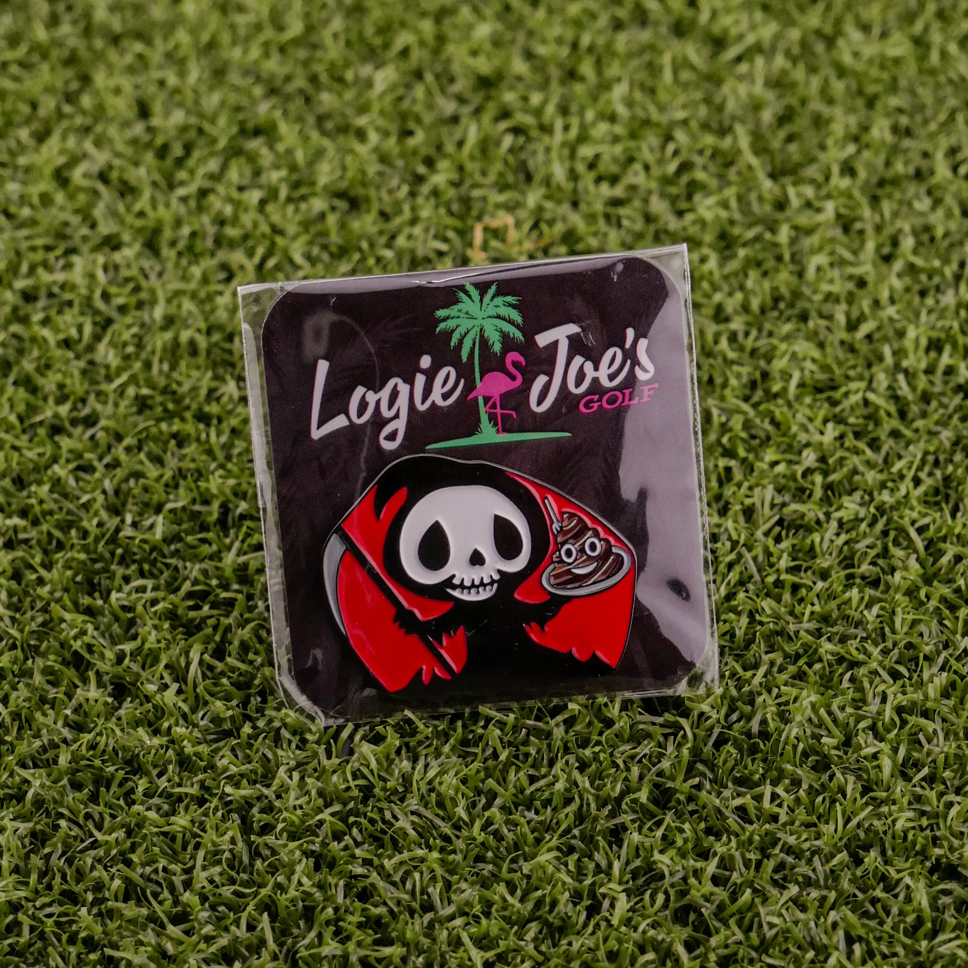 A premium Eat Sh*t & Die ball marker pin by Ballmarkers by Logie Joes, featuring a cartoon grim reaper with a golf club and Logie Joe’s Golf in white and pink, displayed on artificial green grass.
