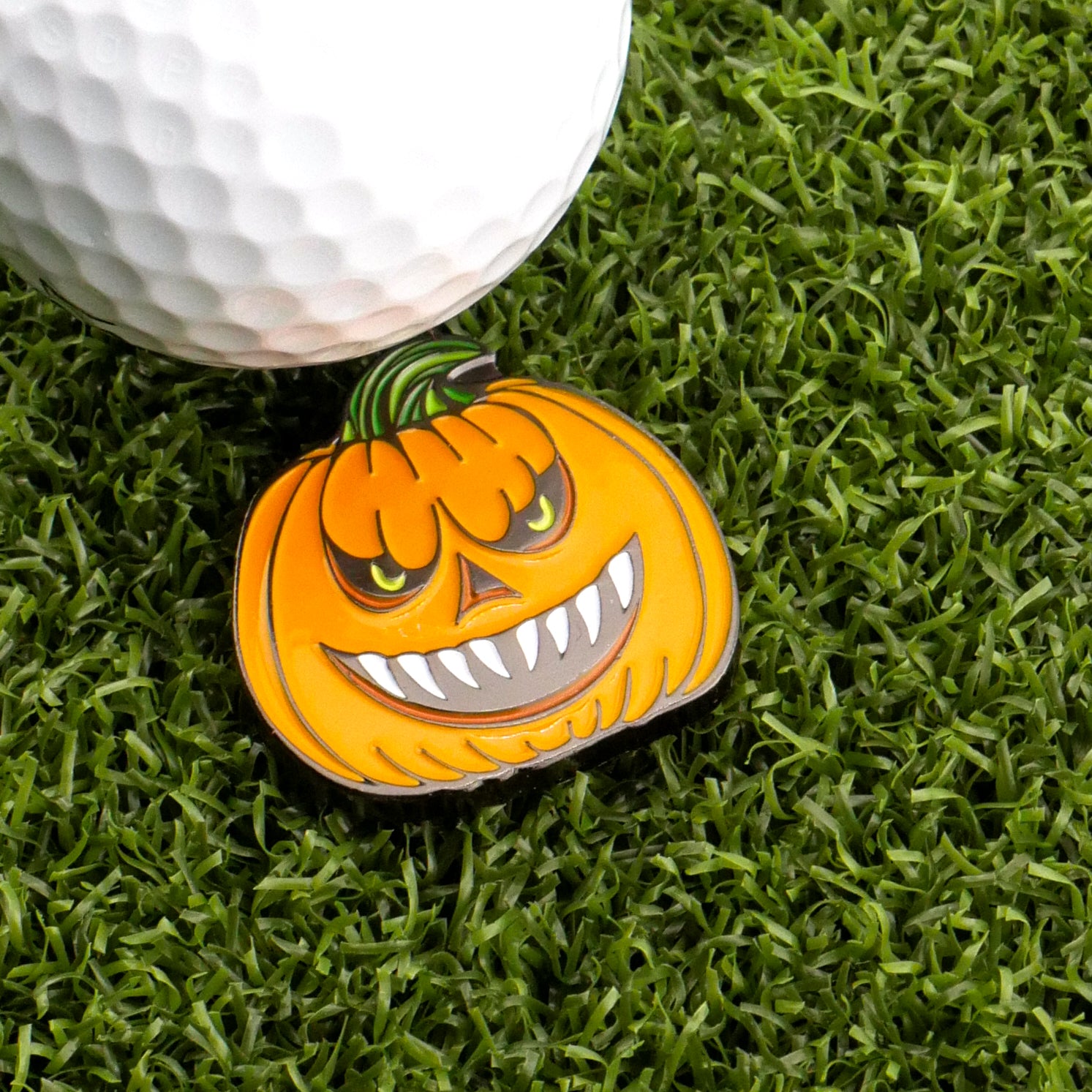 A golf ball sits on artificial grass next to Logie Joe's Golf Evil Jack-O-Lantern Ball Marker, shaped like a grinning, menacing pumpkin with sharp teeth.