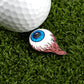 A white golf ball sits on green grass beside Ballmarkers.com's Eye Ball golf ball marker, which is shaped like a blue eyeball with red flame-like streaks.