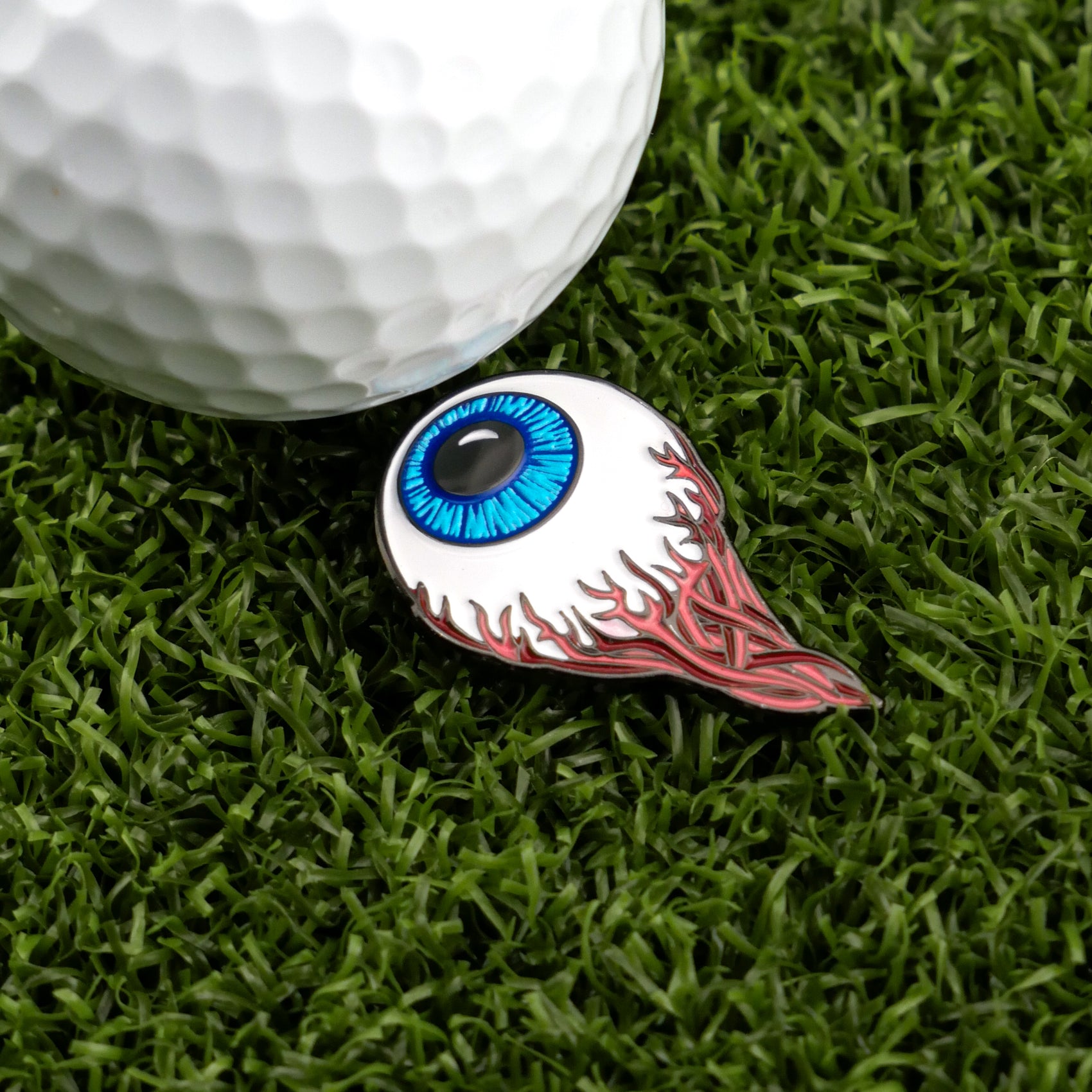 A white golf ball sits on green grass beside Ballmarkers.com's Eye Ball golf ball marker, which is shaped like a blue eyeball with red flame-like streaks.