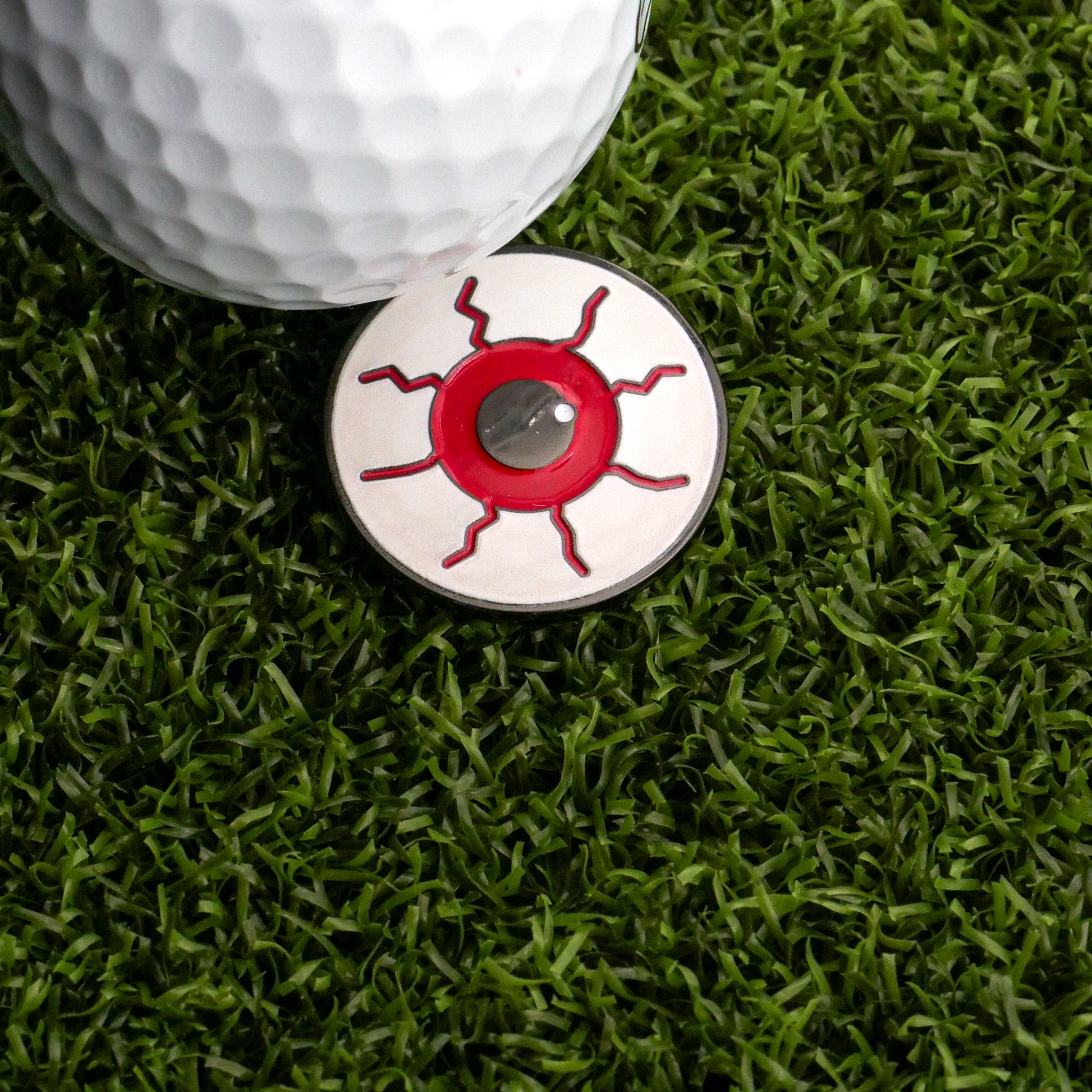 A golf ball sits on artificial putting green turf beside the I See Eyeball Ball Marker by Ballmarkers.com, a distinctive golf accessory with a red sun-like pattern and a silver center.
