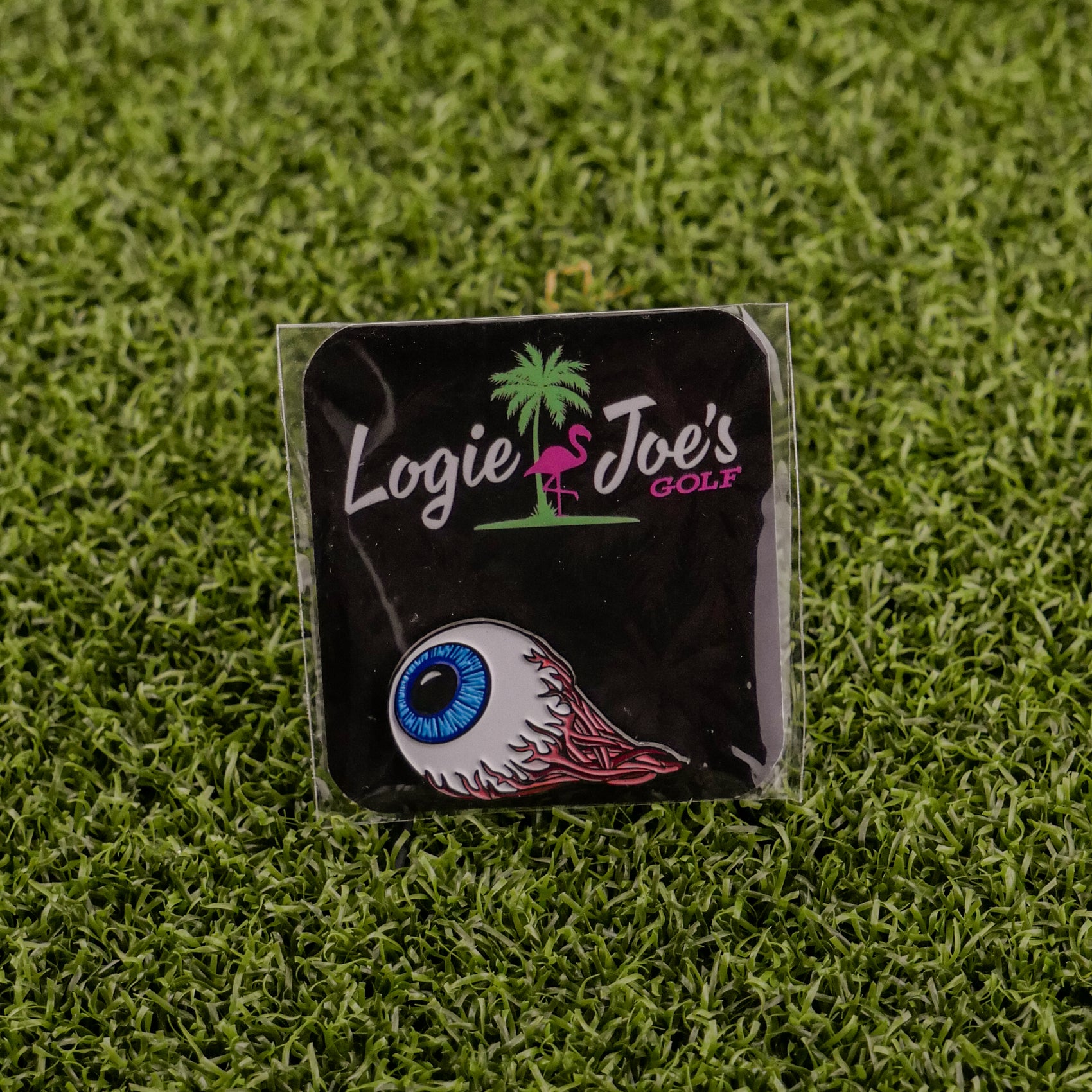 The Eye Ball pin from Ballmarkers by Logie Joes features a blue eyeball with red flames on artificial grass, attached to a branded card with a palm tree and flamingo—an eye-catching golf accessory for your collection.