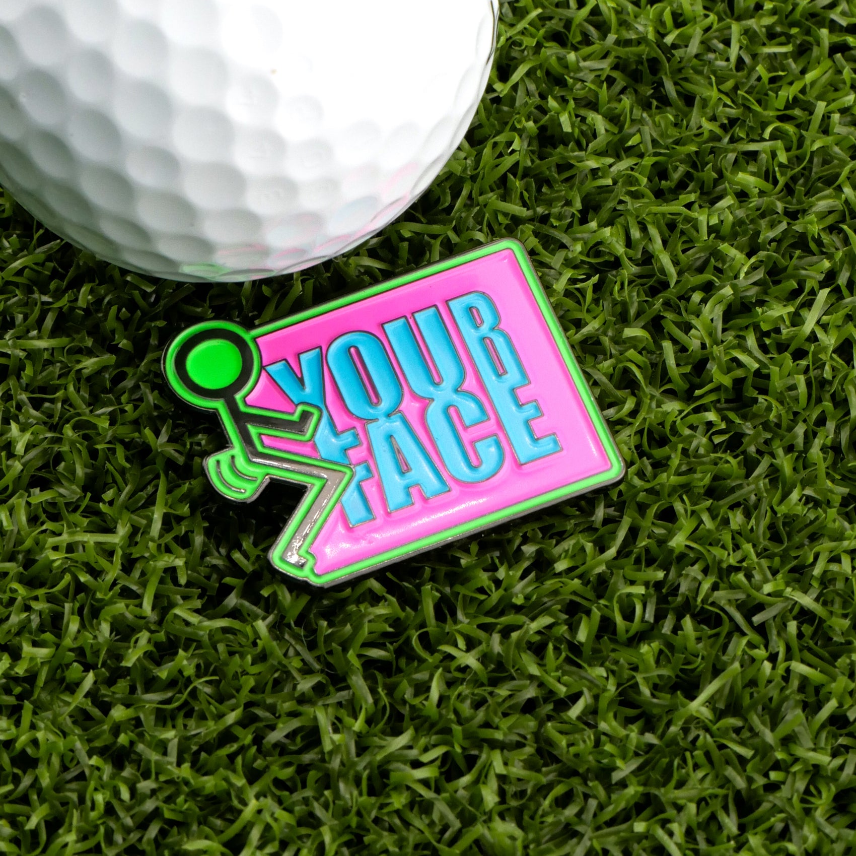 A golf ball and the “F” Your Face Ball Marker by Logie Joe's Golf sit on green grass. The colorful pin features a stick figure and bold blue “YOUR FACE” text on a pink background, adding humor to your golf accessories.