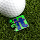 A golf ball sits on artificial grass next to Logie Joe's Golf "F" It premium ball marker, which features a green stick figure kicking the word "it" in bold blue—a humorous gift for golf lovers.