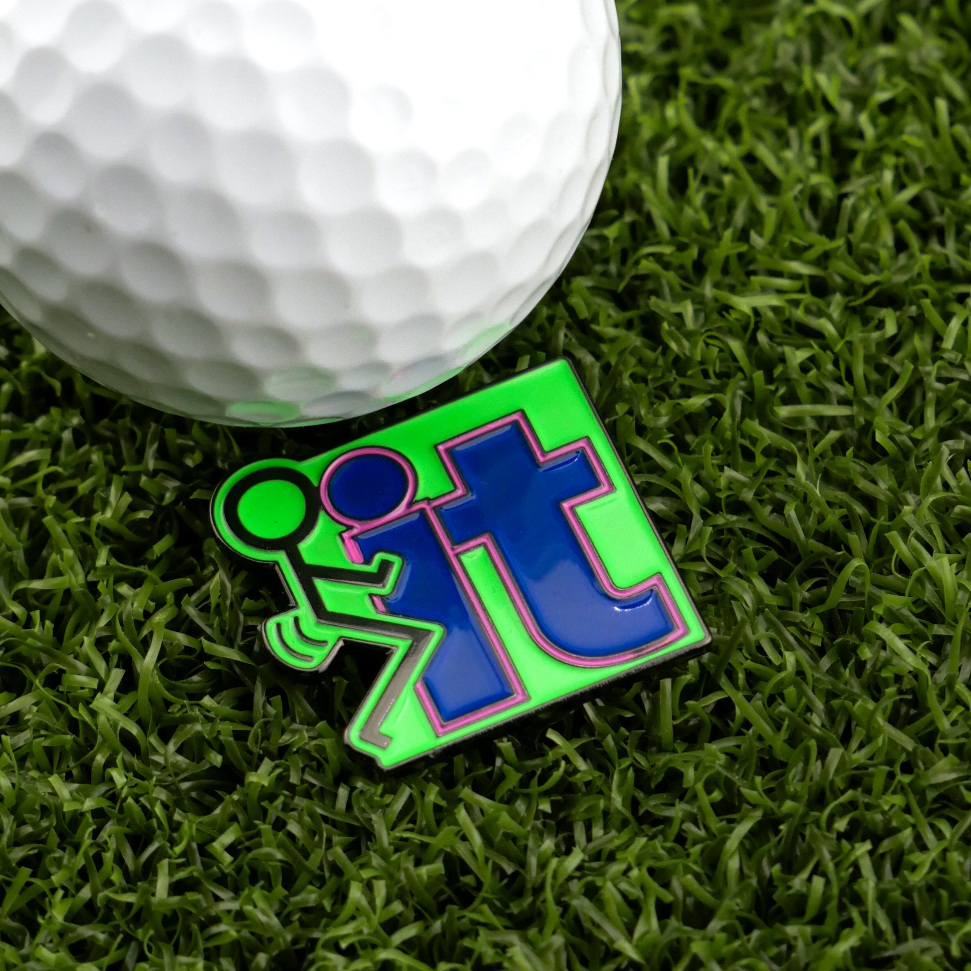 A golf ball sits on artificial grass next to Logie Joe's Golf "F" It premium ball marker, which features a green stick figure kicking the word "it" in bold blue—a humorous gift for golf lovers.