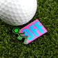 A golf ball on artificial green grass sits beside Logie Joe's Golf “F” Off ball marker, featuring a green stick figure pushing OFF in bold blue letters on a pink background—perfect for golfers seeking fun, premium accessories.