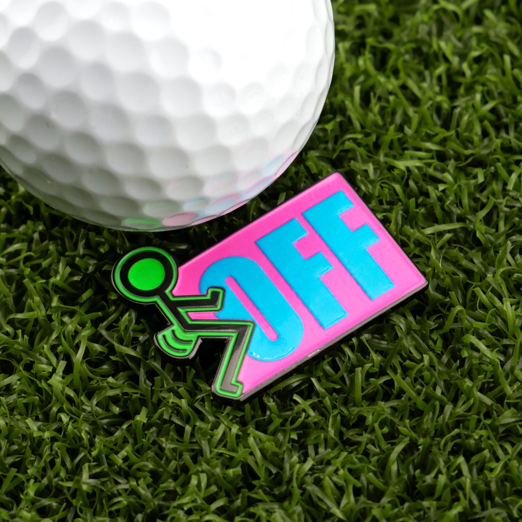 A golf ball on artificial green grass sits beside Logie Joe's Golf “F” Off ball marker, featuring a green stick figure pushing OFF in bold blue letters on a pink background—perfect for golfers seeking fun, premium accessories.