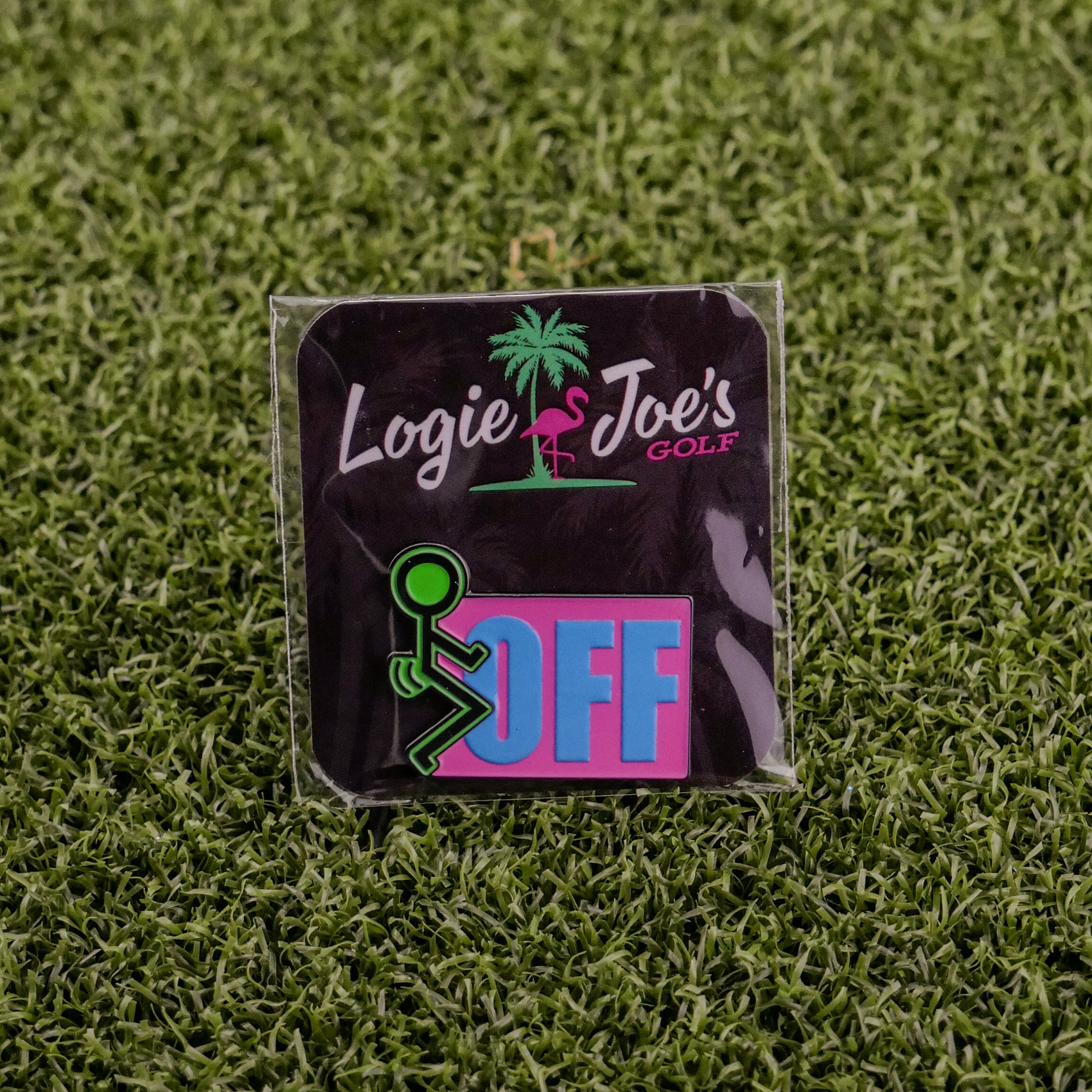 The “F” Off ball marker by Logie Joe's Golf displays pink, green, and white branding above a palm tree, plus a green stick figure beside the blue and pink word OFF—an eye-catching, humorous marker shown on artificial grass.