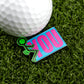 A golf ball on artificial grass sits next to Logie Joe's Golf “F” You marker—a colorful accessory featuring a green stick figure and the word YOU in large pink letters on a blue background.