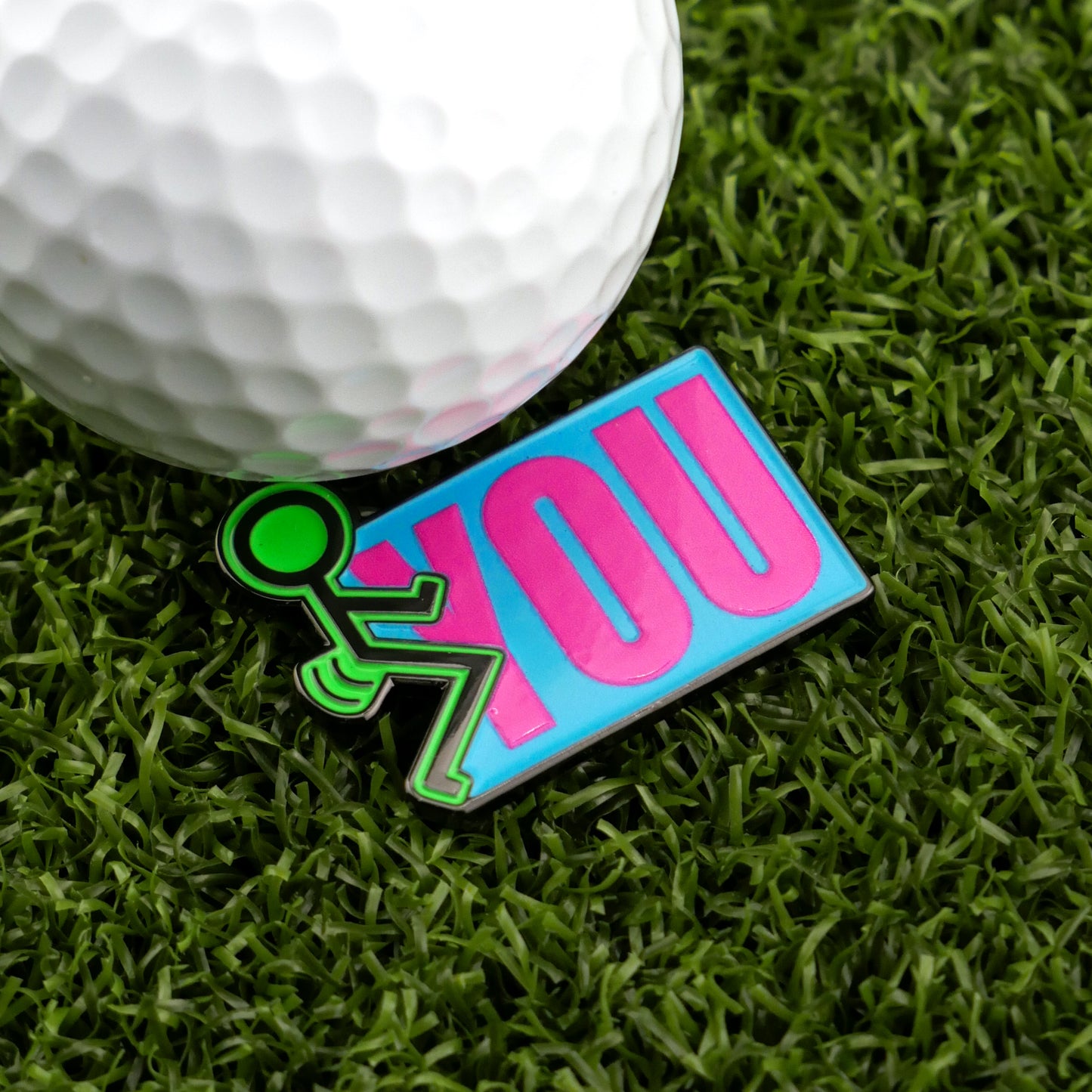 A golf ball on artificial grass sits next to Logie Joe's Golf “F” You marker—a colorful accessory featuring a green stick figure and the word YOU in large pink letters on a blue background.