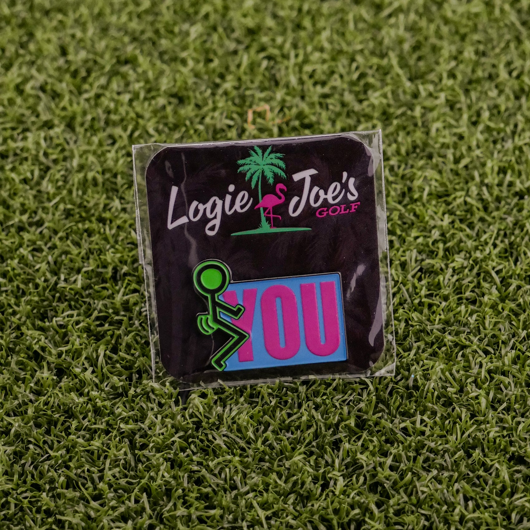 A square golf ball marker on artificial grass shows “F” You with a stick figure, palm tree, and flamingo. Great for your Ballmarkers by Logie Joes collection.