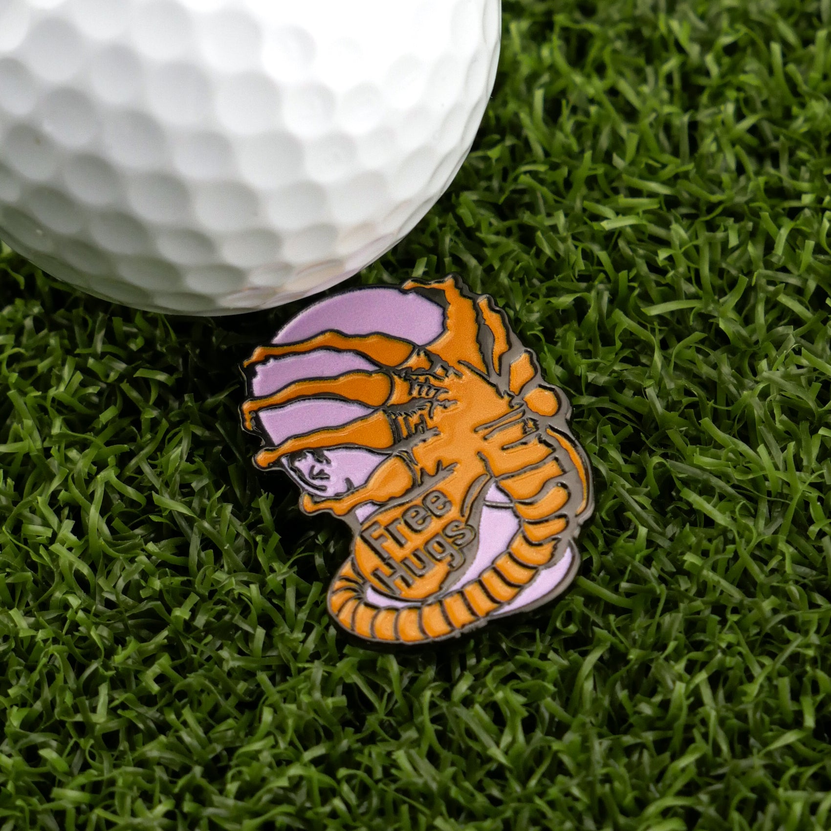 A golf ball rests on artificial grass beside the Face Hugger by Ballmarkers.com—a premium ball marker shaped like an alien embracing a human face with "Free Hugs" inscribed, making it a truly unique golf accessory.