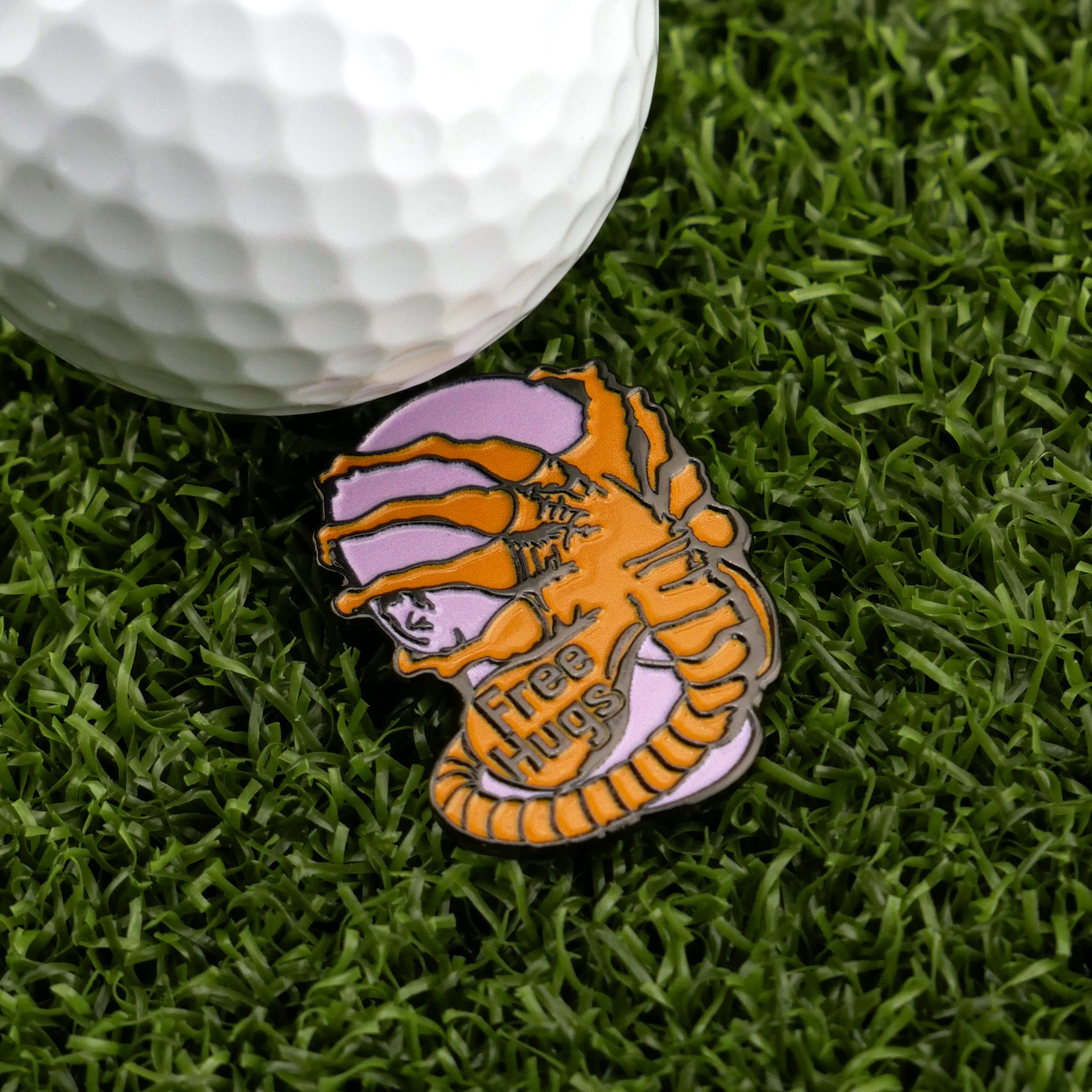 A golf ball rests on artificial grass beside the Face Hugger by Ballmarkers.com—a premium ball marker shaped like an alien embracing a human face with "Free Hugs" inscribed, making it a truly unique golf accessory.