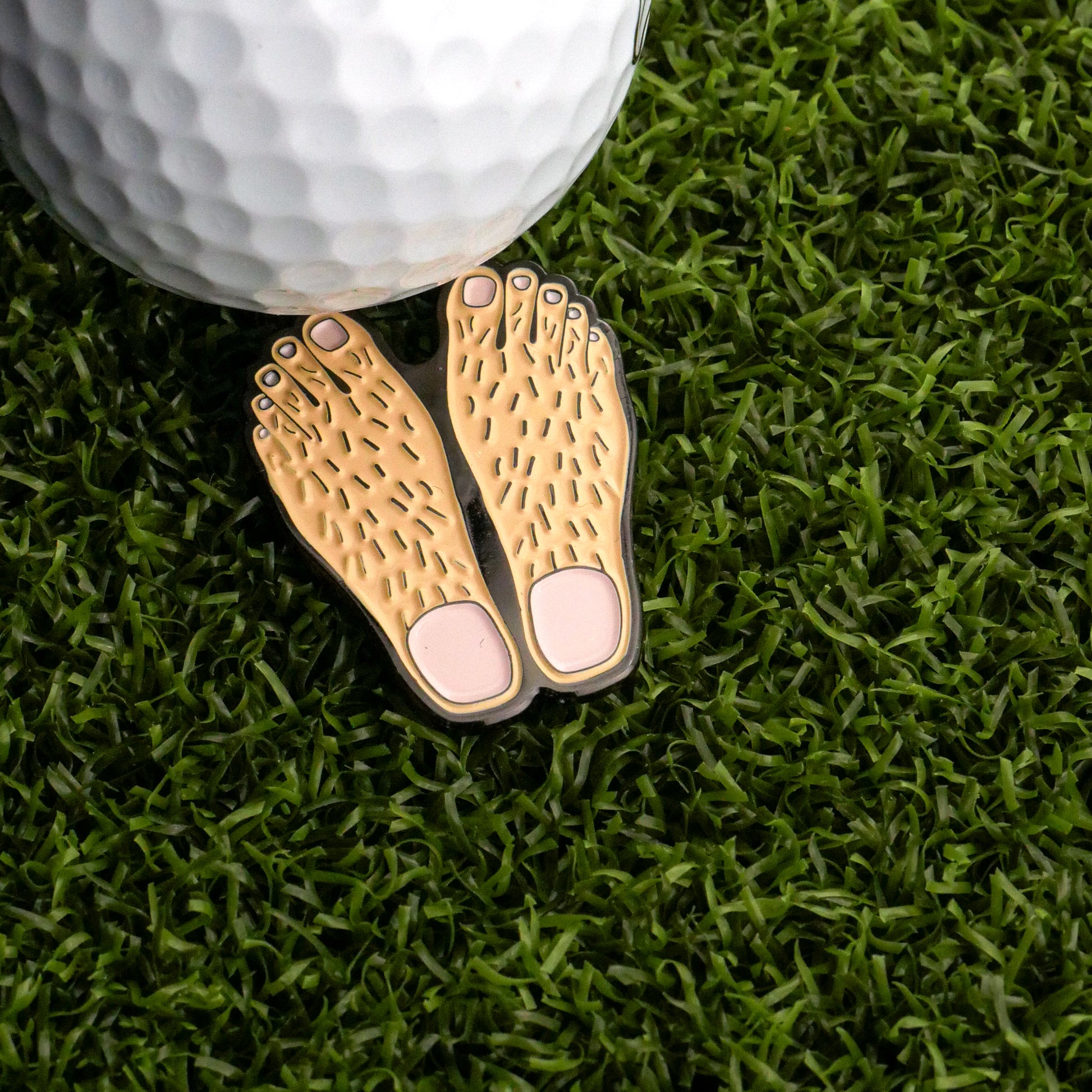 A golf ball rests on artificial grass next to the Logie Joe's Golf Feet Ball Marker, a fun golfer accessory shaped like cartoonish, hairy bare feet.