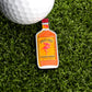 A golf ball on green grass sits beside the Cinnamon Whiskey Ball Marker by Logie Joe's Golf, featuring a pink flamingo illustration—an eye-catching addition to your golf accessories.