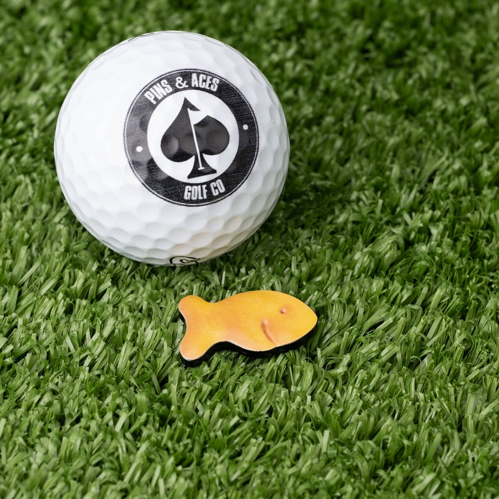 A white golf ball with a Pins and Aces logo rests on green turf beside a yellow fish-shaped candy and the stylish Fish Cracker Ball Marker by Pins and Aces.