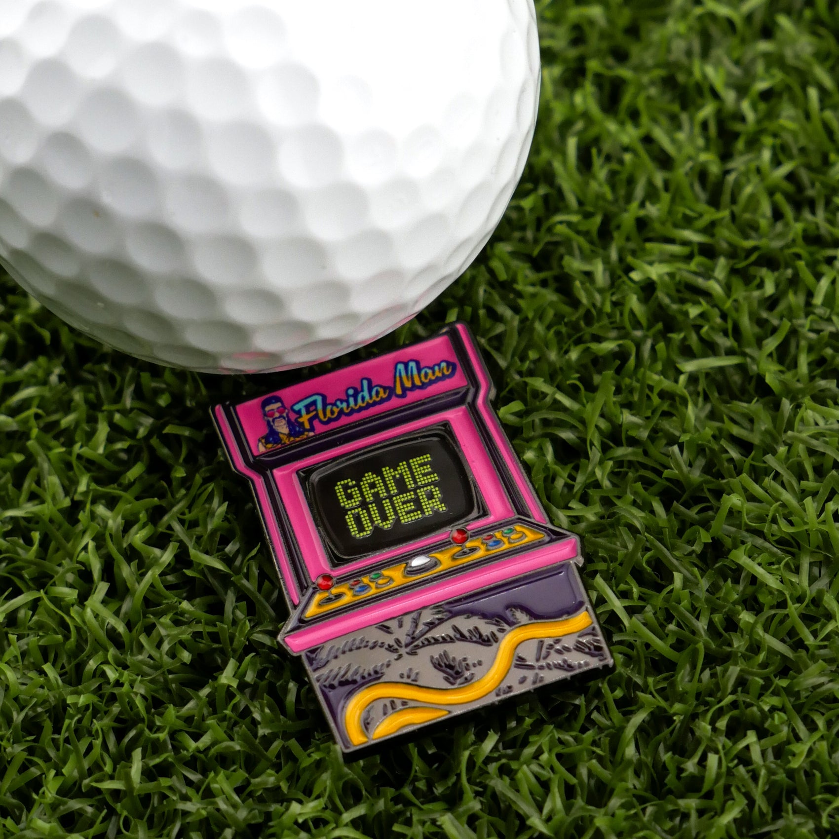 A Logie Joe's Golf ball with a premium marker sits on artificial grass next to a Florida Man (Game Over) arcade pin, featuring "GAME OVER" on the screen.