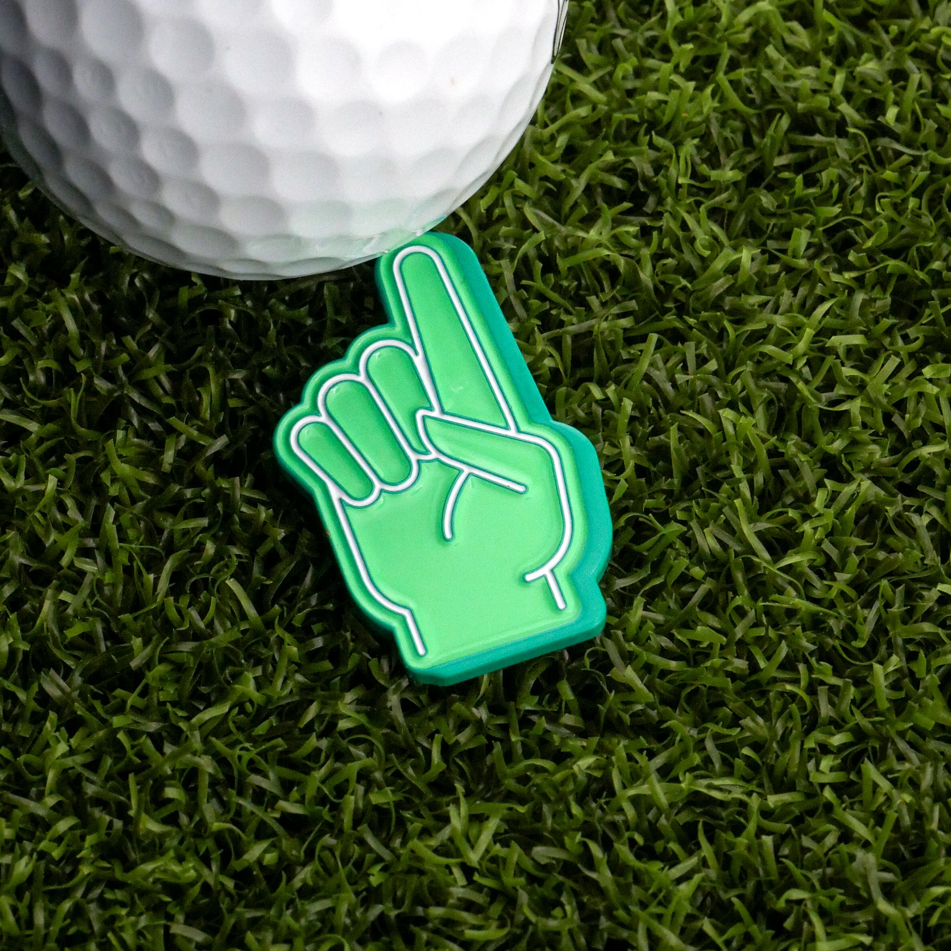 A Foam Finger #1 Ball Marker by Ballmarkers.com and a golf ball rest on artificial green grass, the foam finger pointing upward in celebration.