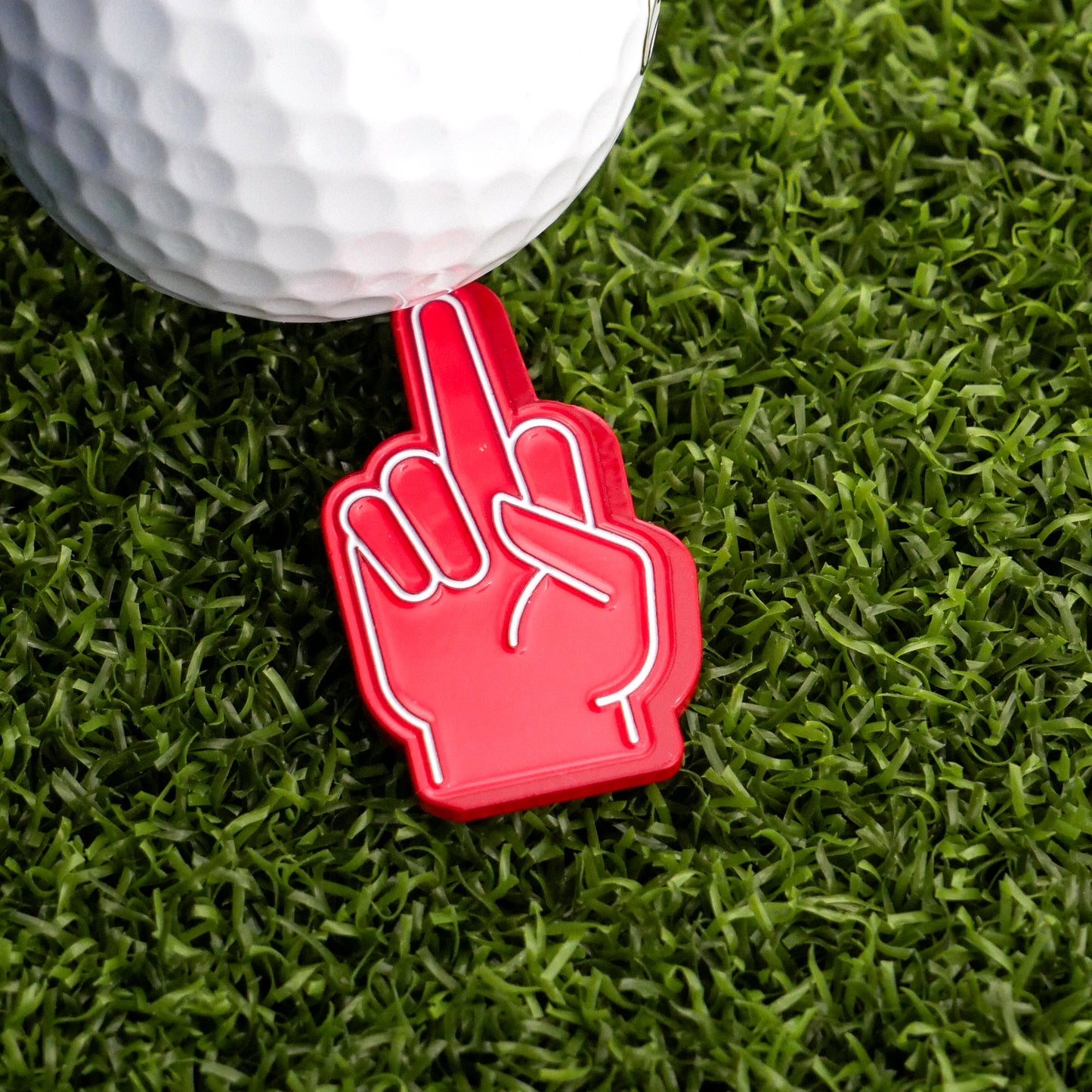 The Ballmarkers.com Foam Finger Middle Finger Ball Marker, a red plastic hand-shaped marker, is seen on green artificial grass beside a golf ball—perfect for expressing golf frustration.