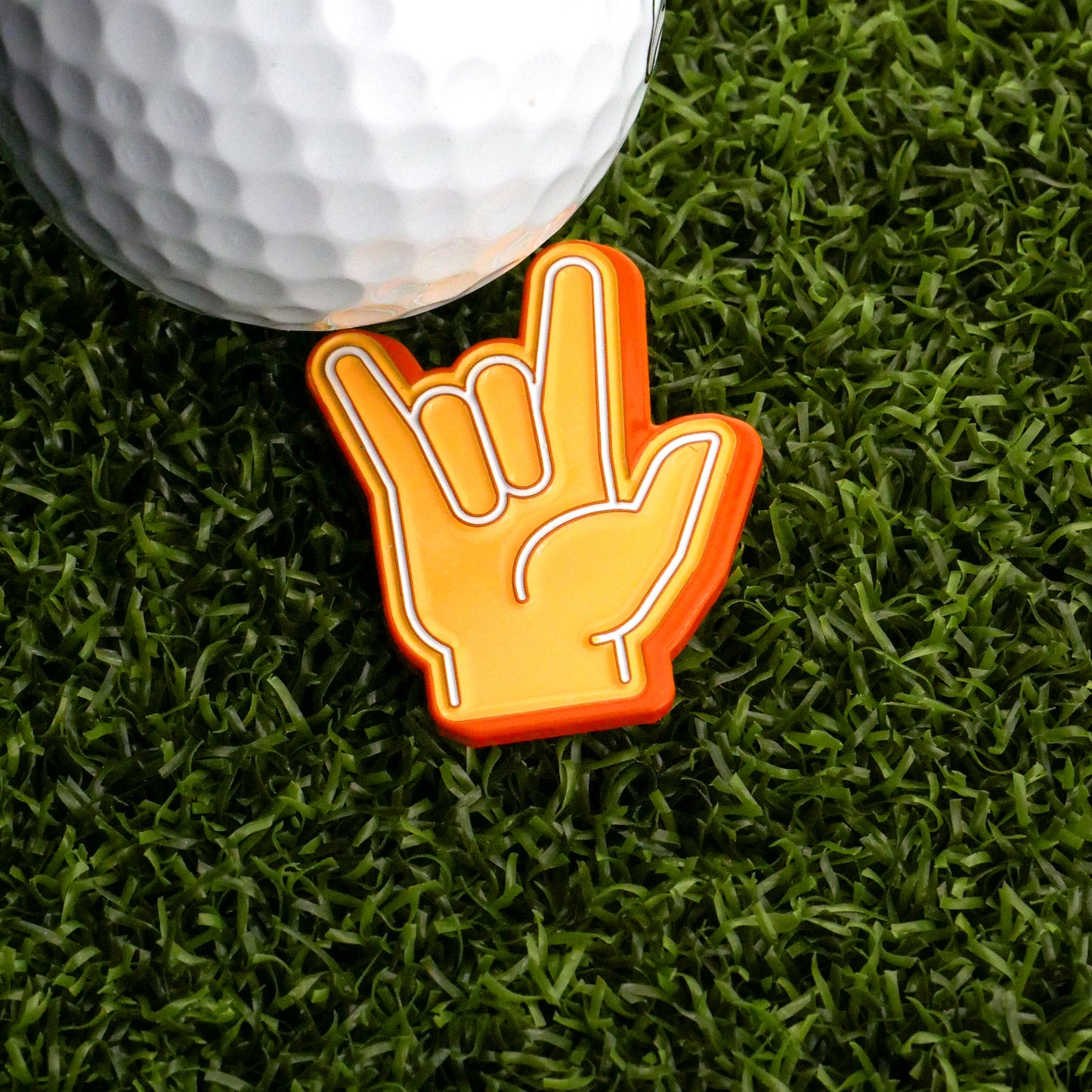 A golf ball sits on artificial grass beside the Ballmarkers.com Foam Finger Rock On Ball Marker, shaped like a hand making the rock on gesture with vibrant orange and yellow accents.