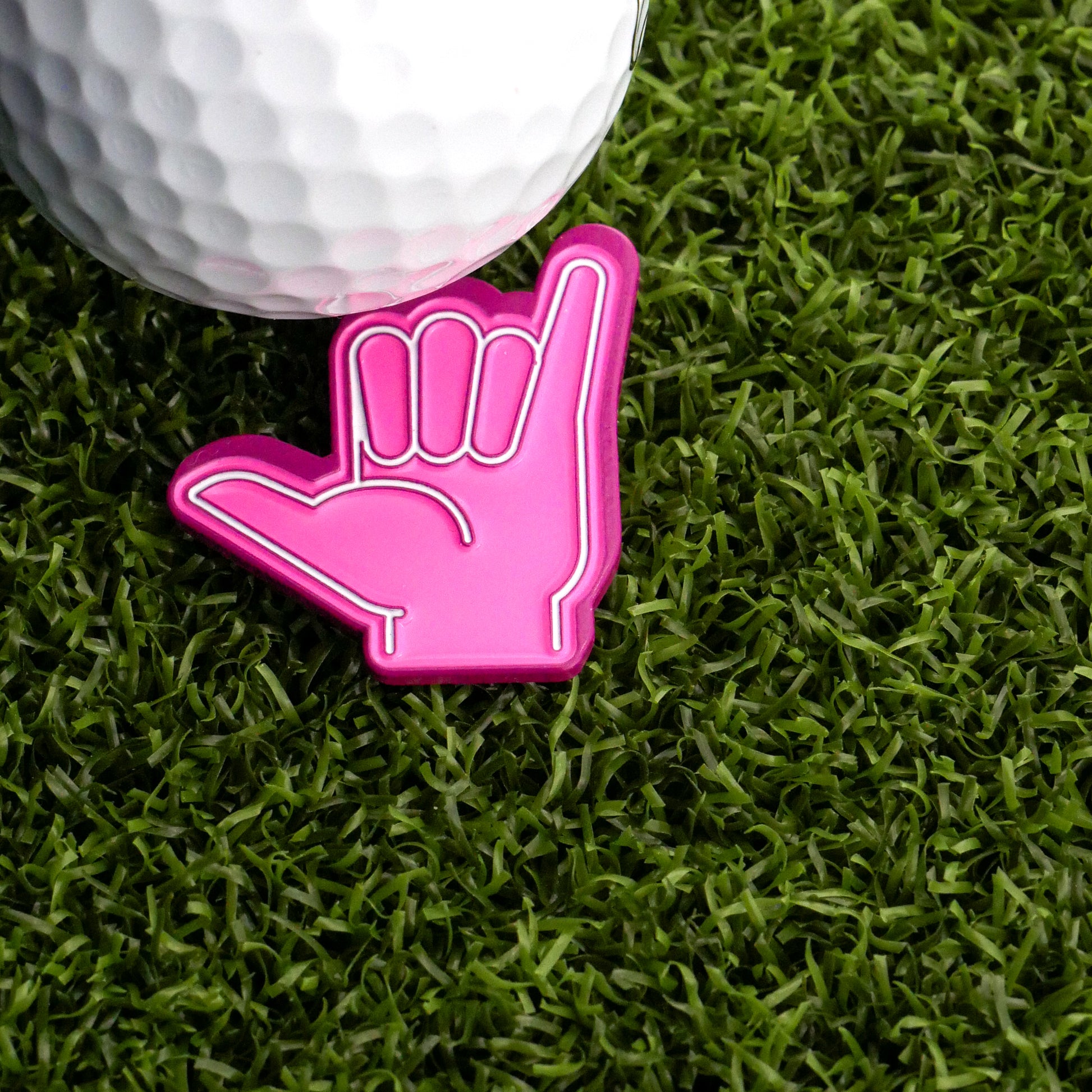 A white golf ball rests on green turf next to the Ballmarkers.com Foam Finger Shaka Ball Marker, shaped like a pink "hang loose" hand—adding fun and flair to your short game.