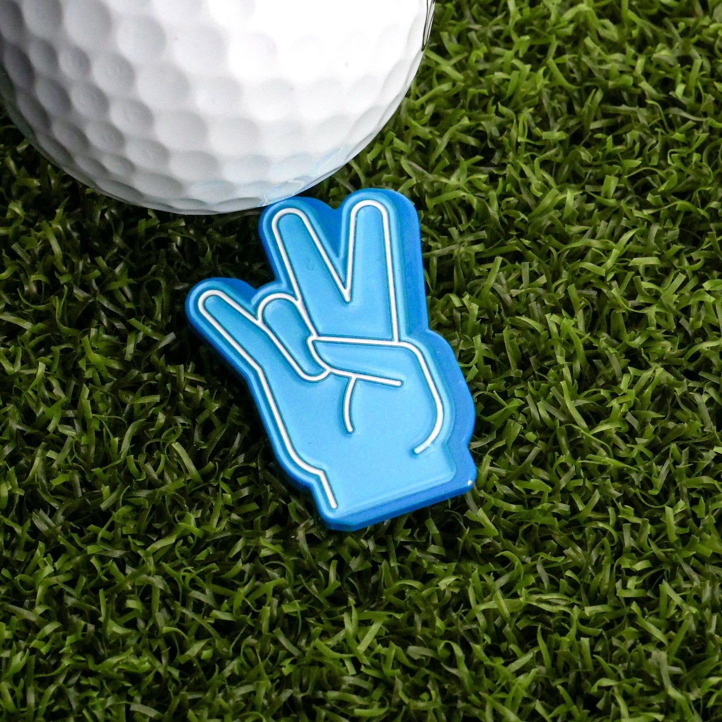 The Foam Finger Shocker Ball Marker by Ballmarkers.com—a blue plastic hand—rests on green turf beside a white golf ball, offering a playful way to mark your spot.