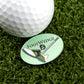 A golf ball on artificial grass sits next to a green oval Foot Wedge golf ball marker by Logie Joe's Golf, featuring a golf shoe and club design. Available at Ballmarkers.com.