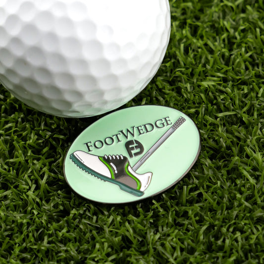A close-up of Logie Joe's Golf Foot Wedge Ball Marker—an oval marker with a golf shoe and wedge design—beside a golf ball on artificial grass, set against a light green background. Find unique markers at Ballmarkers.com.