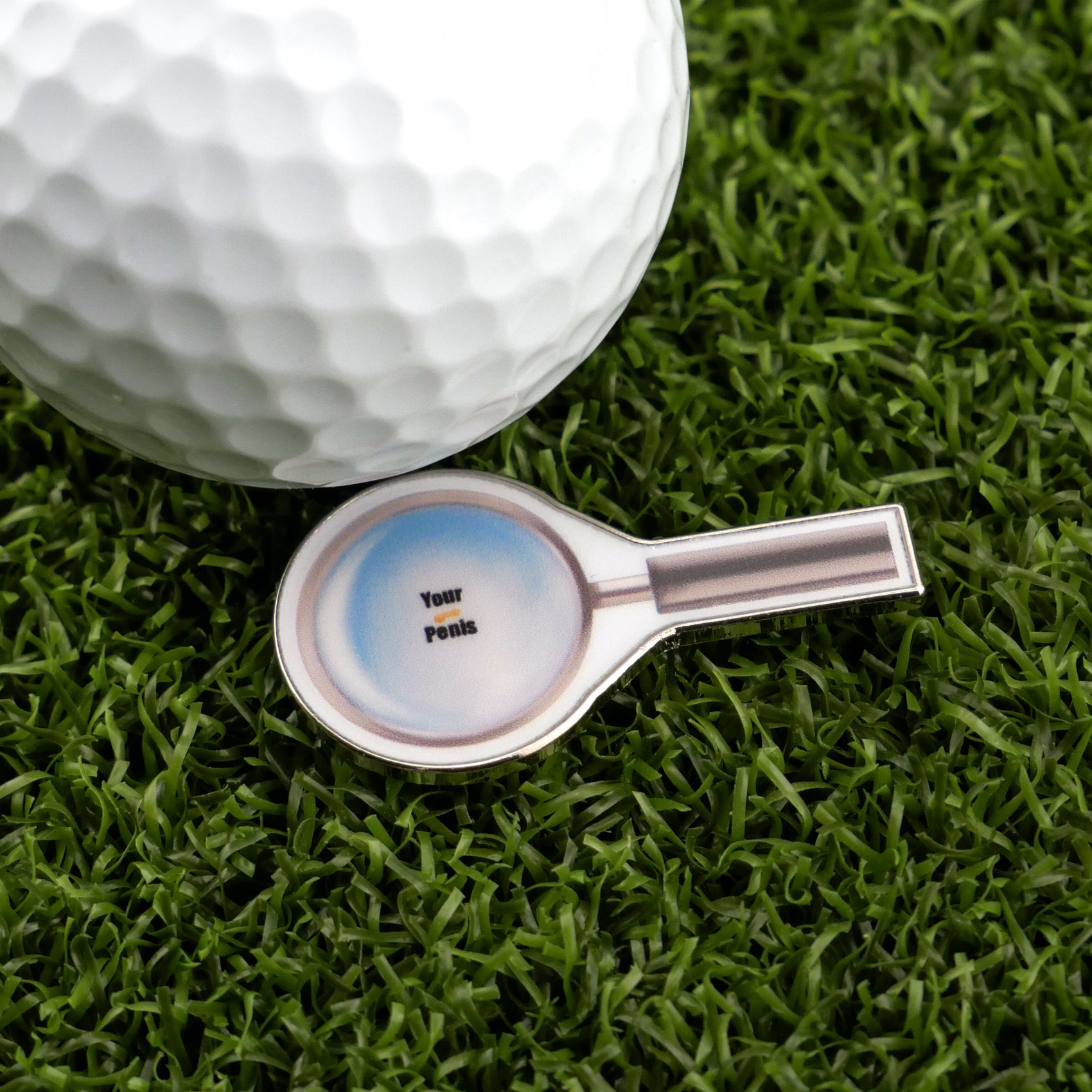 On artificial grass, a golf ball sits by Logie Joe’s Golf "Magnify Glass" premium ball marker—shaped like a magnifying glass with "Your Penis" written in the lens for a humorous touch.