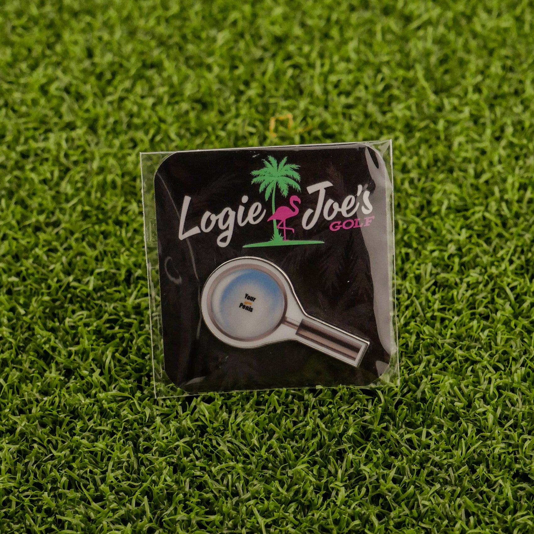 The Logie Joe's Golf "Magnify Glass" pin features a magnifying glass design with "putter" inside, set on green turf—a unique golf ball marker and a funny accessory for your collection.