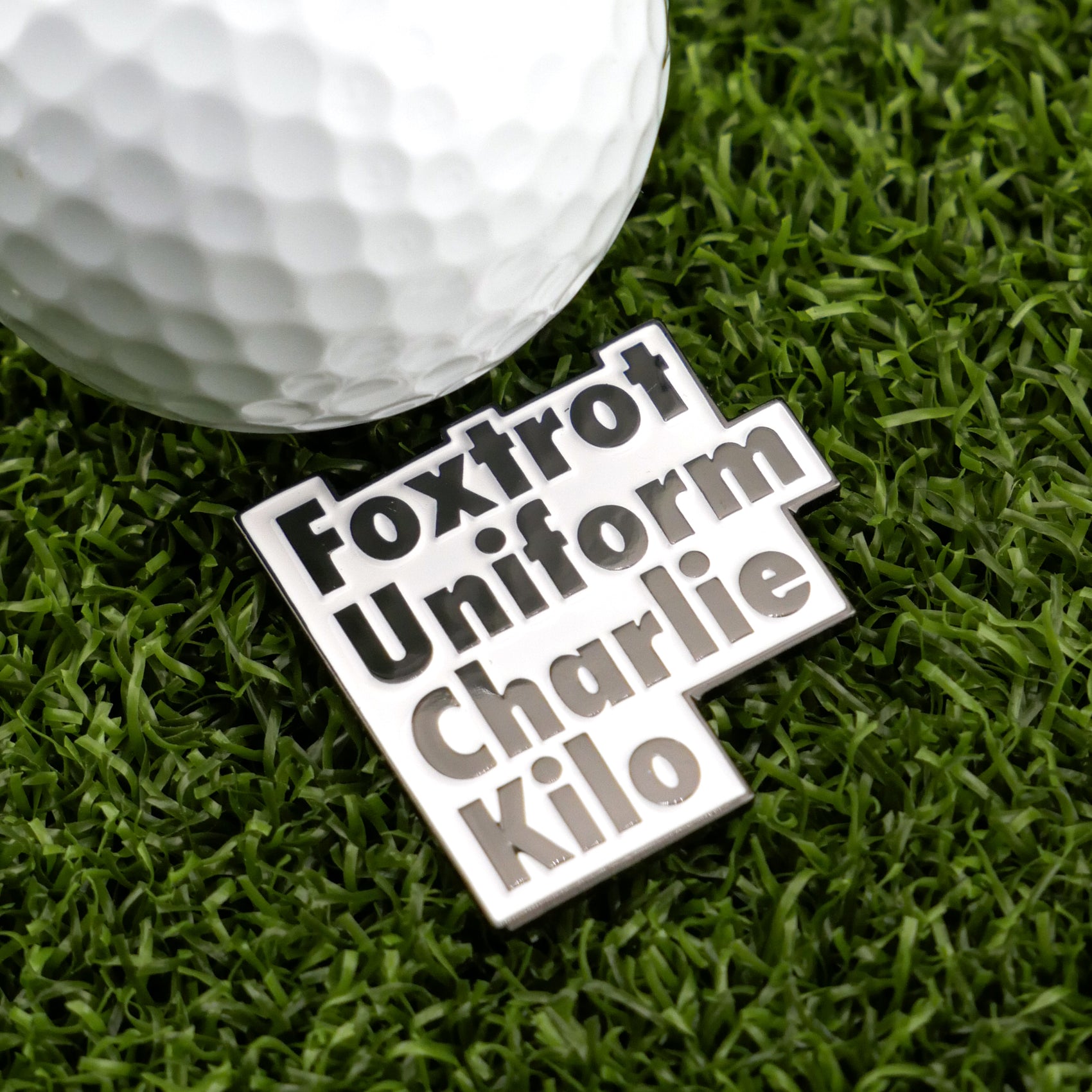 A golf ball on green grass sits beside Logie Joe’s Golf Foxtrot Uniform Charlie Kilo marker, featuring bold black and white letters for a fun accent to your premium golf accessories.