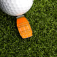A golf ball sits on artificial grass beside the Ballmarkers.com Juice Barrel Ball Marker, an orange, bottle-shaped marker with black and white details, evoking juice boxes from childhood summers.
