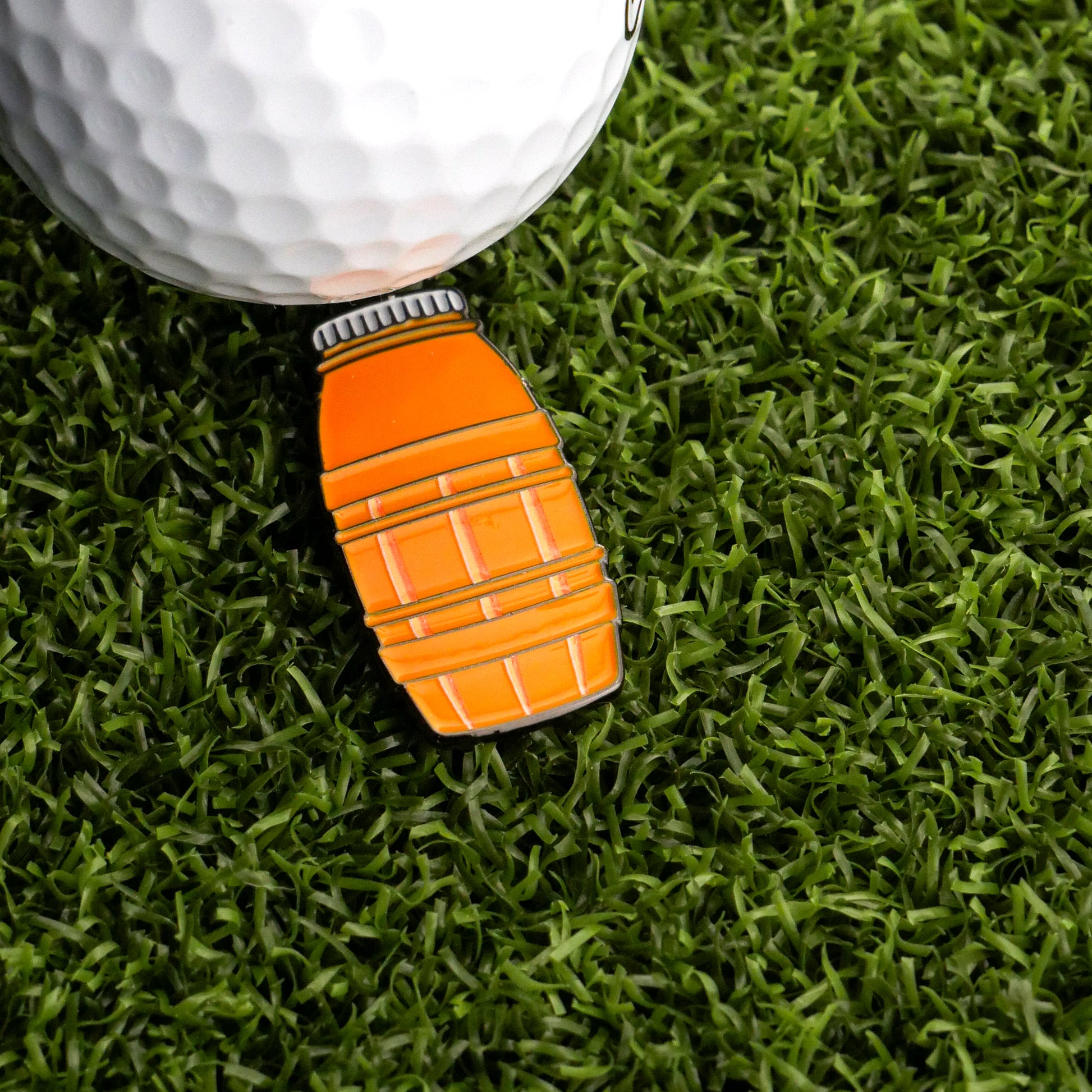 A golf ball sits on artificial grass beside the Ballmarkers.com Juice Barrel Ball Marker, an orange, bottle-shaped marker with black and white details, evoking juice boxes from childhood summers.