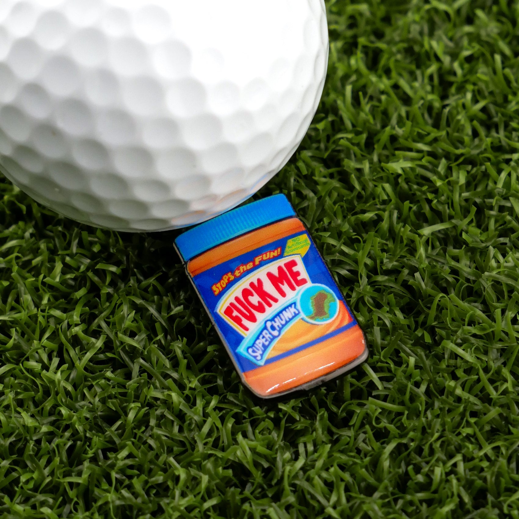 A miniature jar of Ballmarkers.com Chunky Peanut Butter labeled "F*CK ME SUPER CHUNK" rests beside a golf ball and premium golf accessories on artificial green grass.
