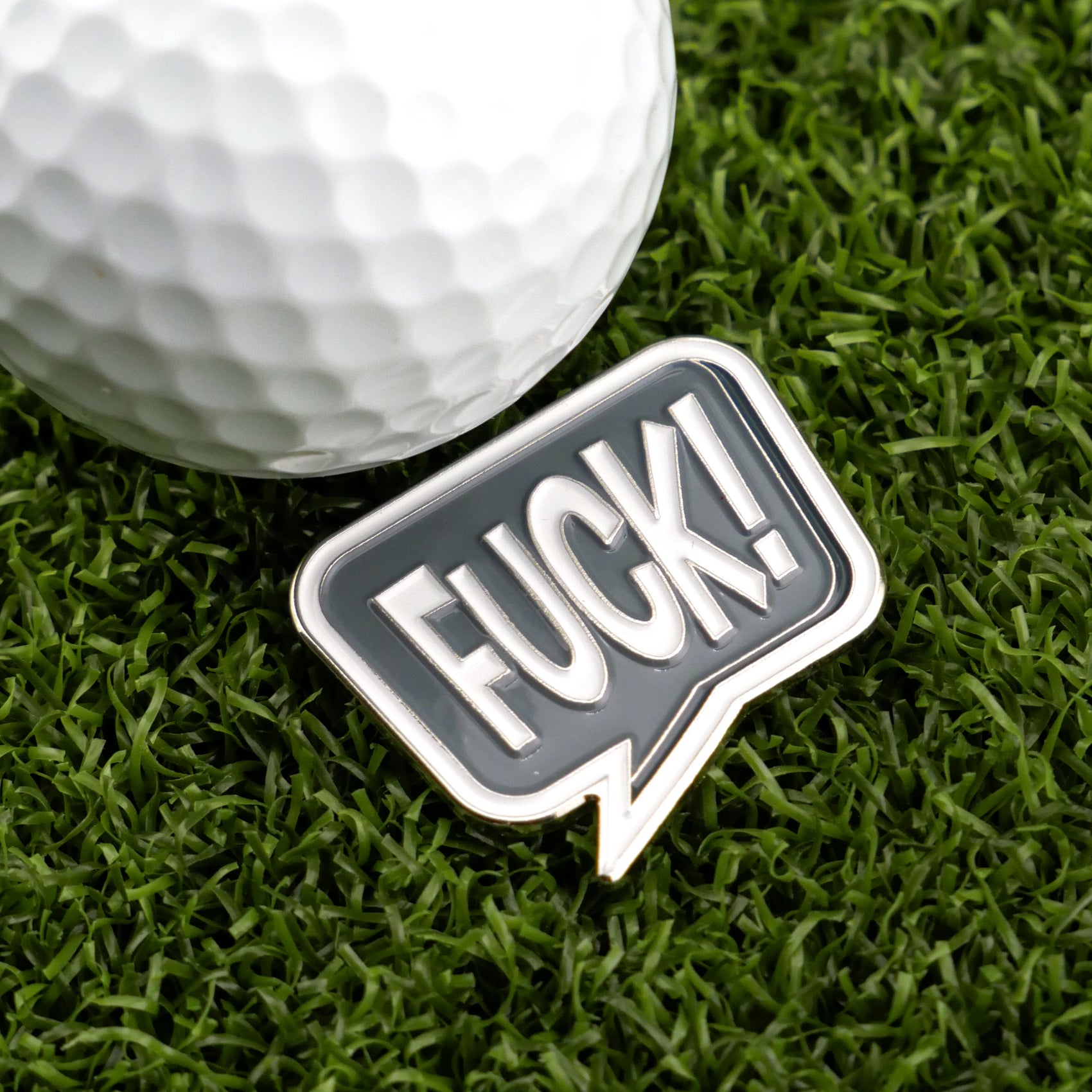 A golf ball sits on artificial grass by the F*ck! Speech Bubble marker from Logie Joe's Golf—a bold, black-and-white accessory perfect for fun-loving golfers seeking a unique way to mark their ball.
