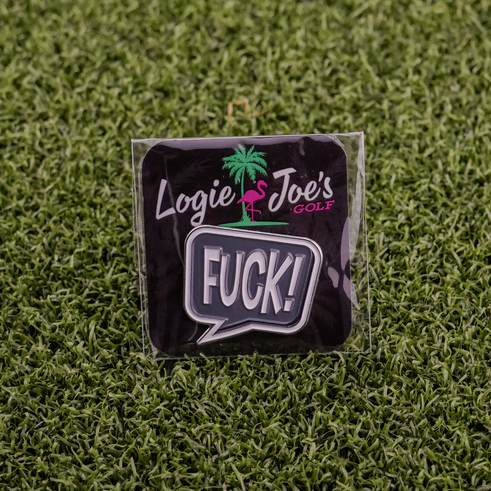 The F*ck! Speech Bubble pin by Logie Joe's Golf features a playful design on branded packaging with a palm tree and flamingo logo, displayed on green turf—ideal for fans of funny golf accessories or unique ball markers.