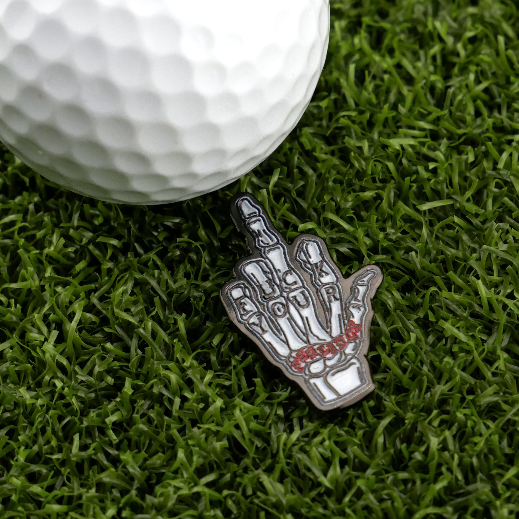 A golf ball rests on artificial grass next to Logie Joe's Golf "F" Your Feelings ball marker—a skeleton hand flipping the bird with SICK PUTT inscribed across its fingers.