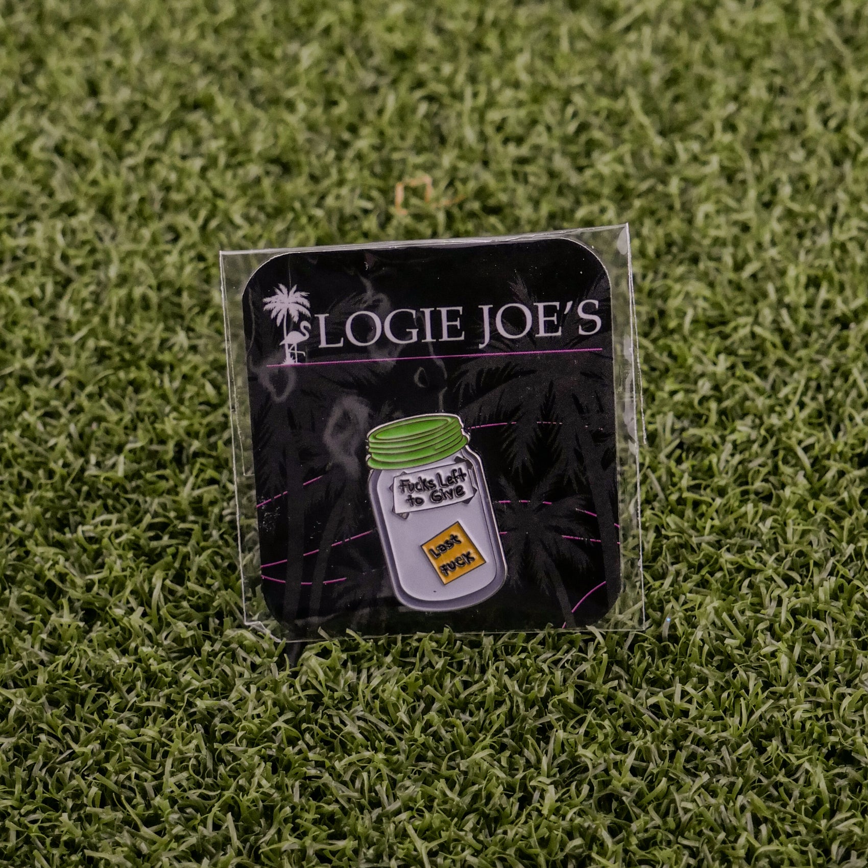The Jar of “F”s enamel pin by Ballmarkers by Logie Joes sits on a branded card atop artificial grass—perfect as a funny ball marker or gift for fans of unique golf accessories.